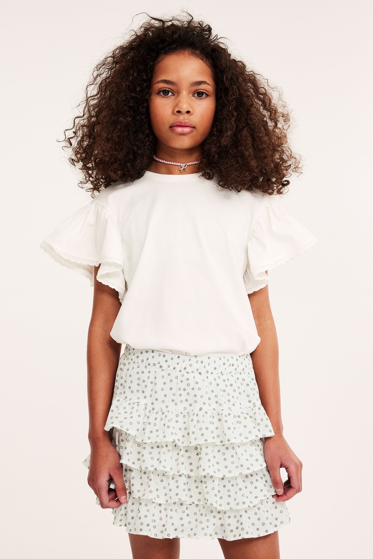 Frontvy av ett barn i offwhite mönstrad volangkjol, offwhite t-shirt och rosa pärlhalsband.