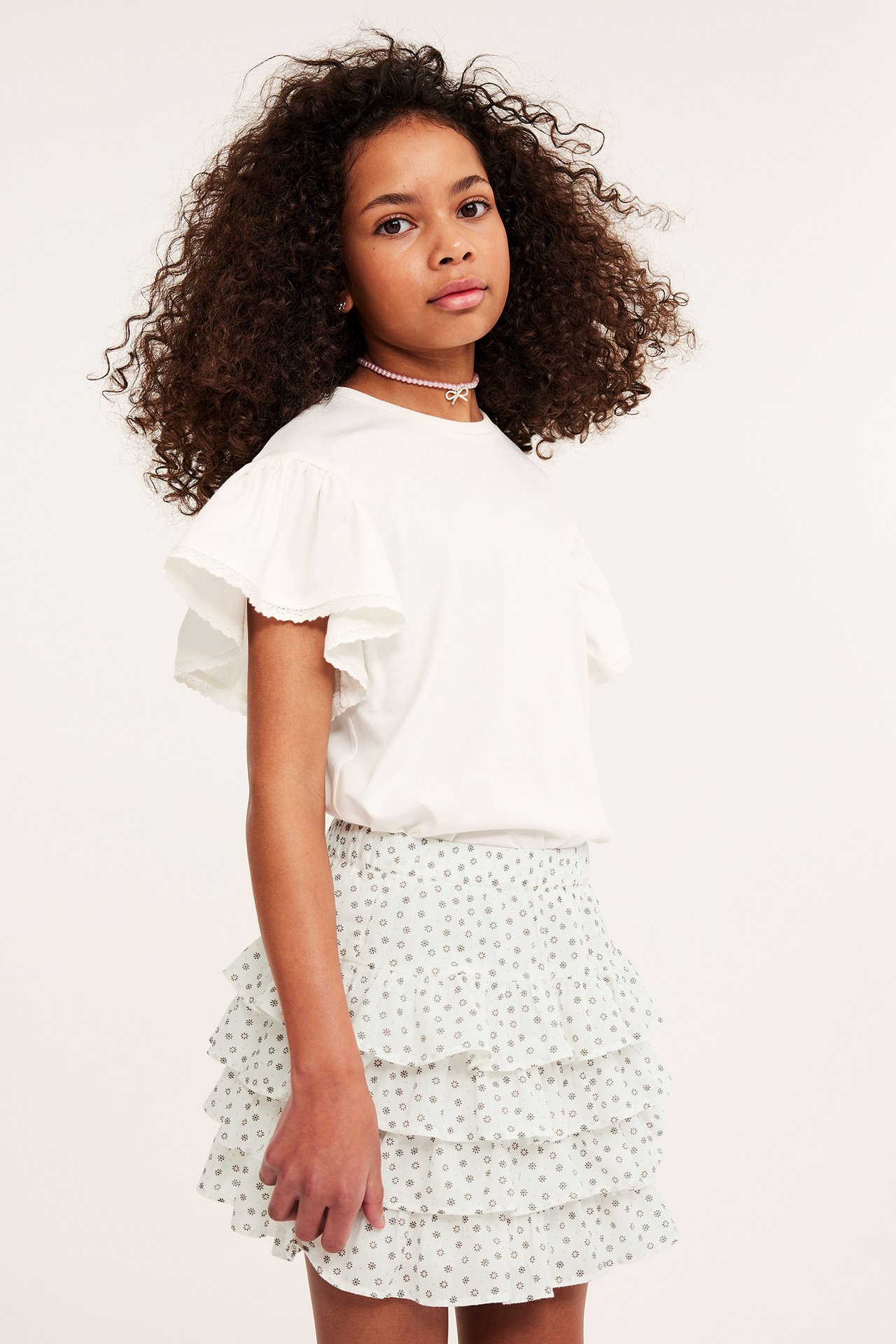 Framifrån: Barn i ljusgrön mönstrad volangkjol, offwhite volang-t-shirt och rosa pärlhalsband med rosett.