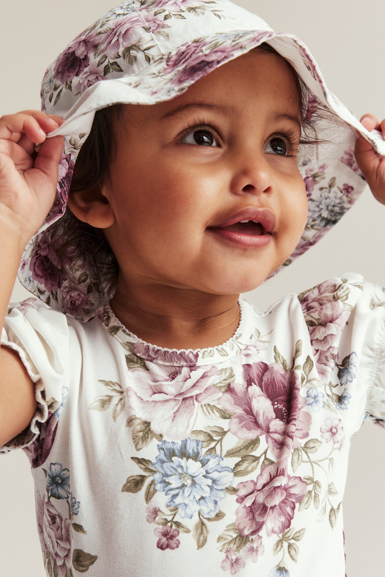 Nærbilde av en baby som har på seg en offwhite blomstret solhatt med bred brem og en matchende blomstret topp.