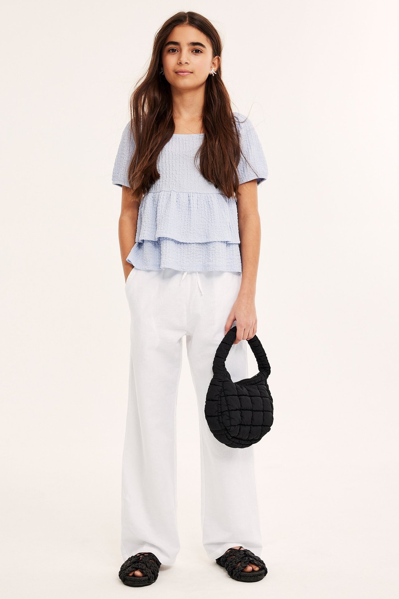 Edestäpäin, lapsi valkoisissa pellavahousuissa, vaaleansinisessä topissa, musta tikattu laukku ja mustat sandaalit.