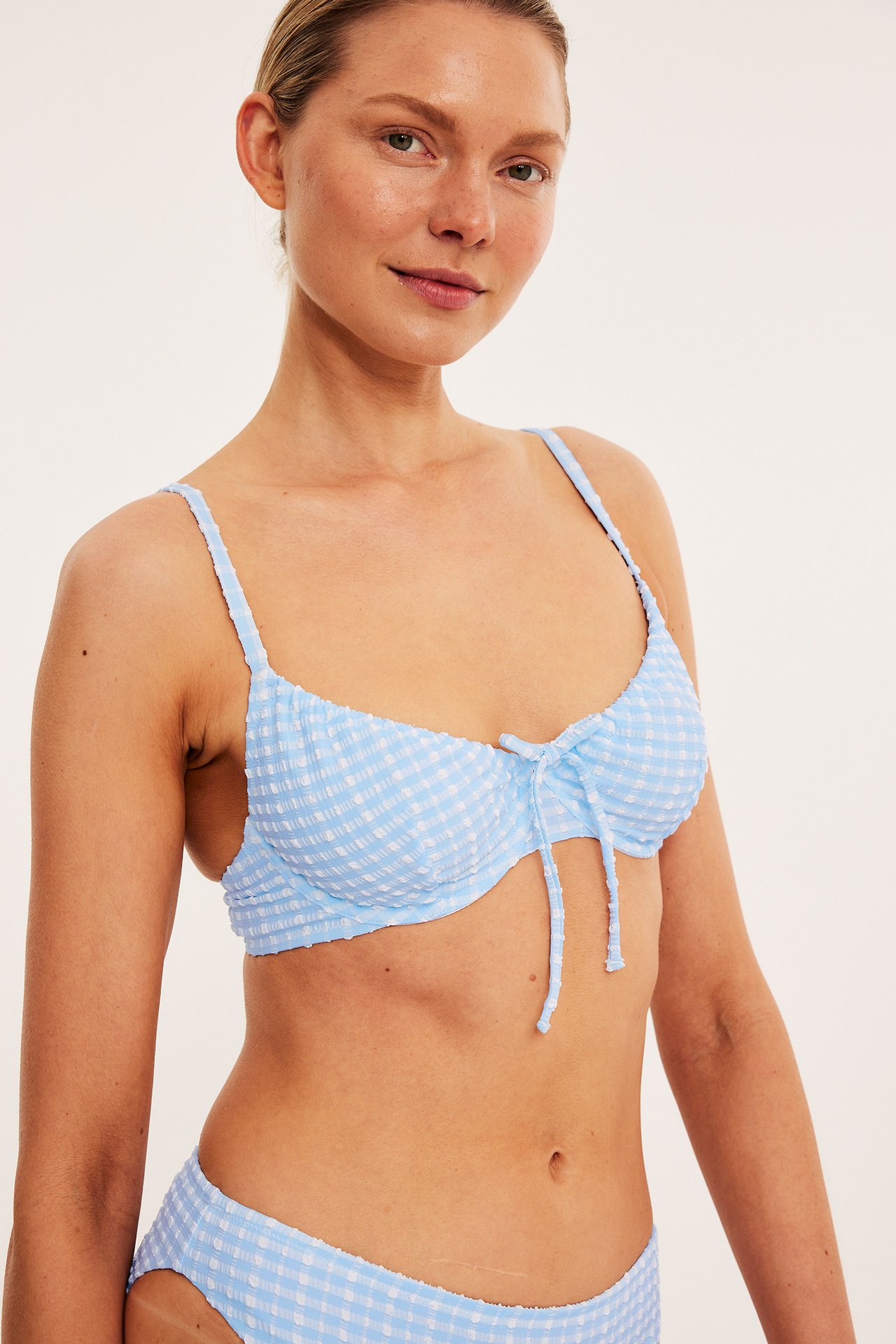 Framifrån, kvinna i ljusblå-vit rutig bikini-bh med knyt framtill och matchande trosa.