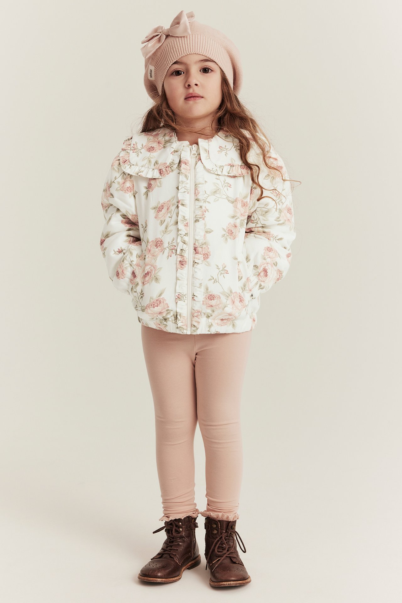 Forfra: Barn i offwhite blomstret bomberjakke med volanger, lyserosa leggings, rosa beret, brune støvler.