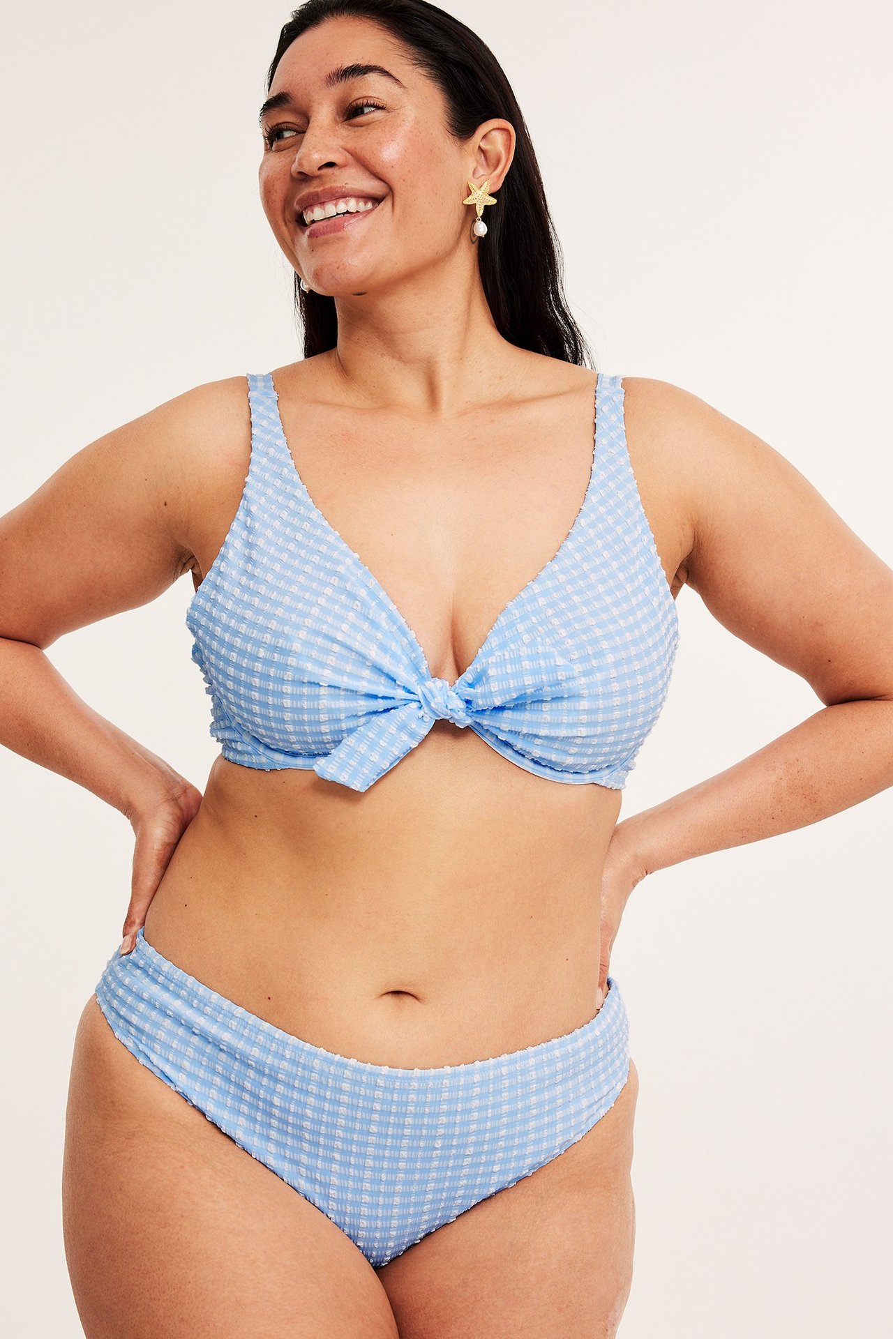 Forfra, kvinne i lyseblå rutete seersucker bikinitruse, matchende topp og sjøstjerneøreringer.