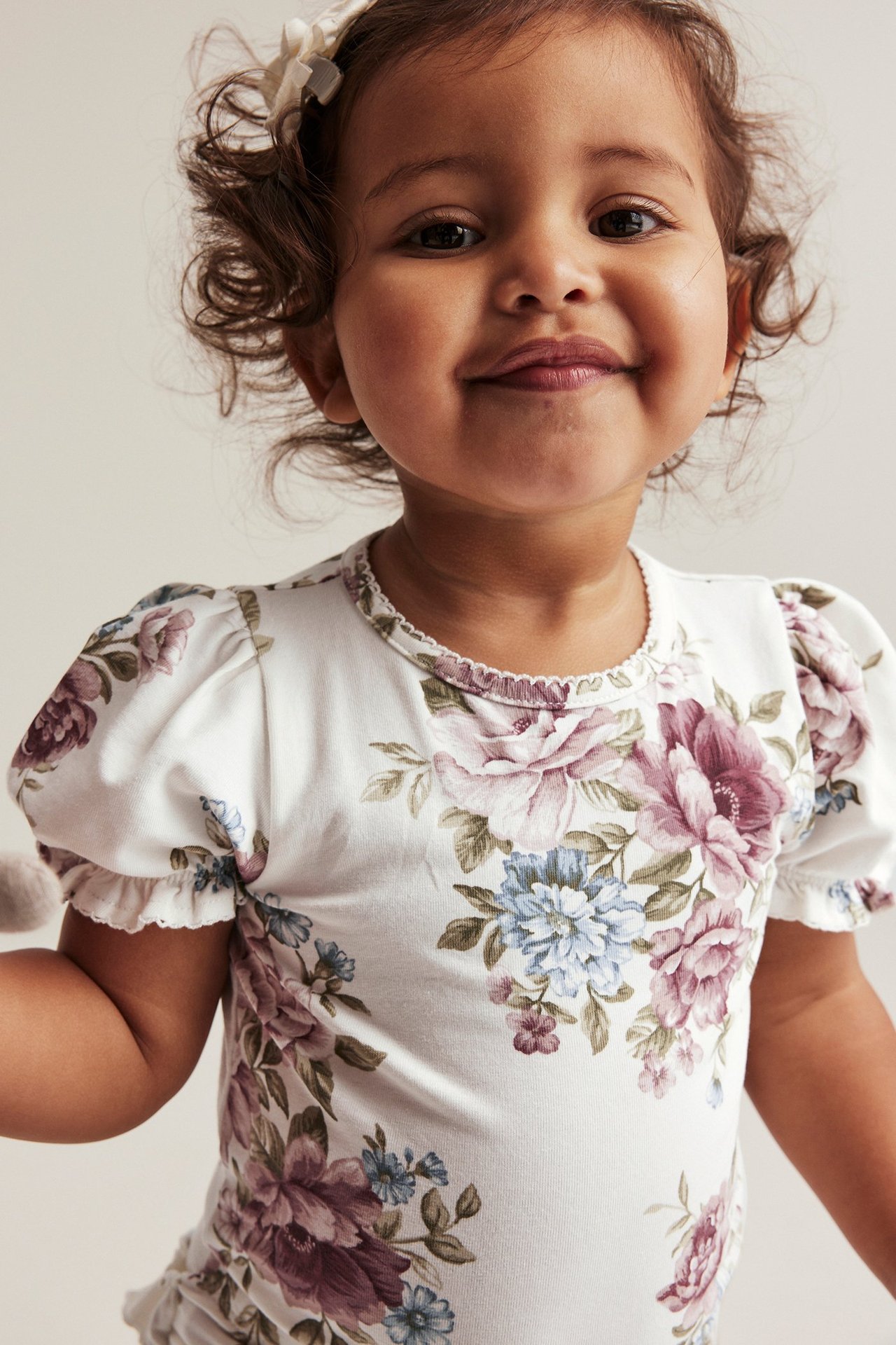 Lähikuva edestä vauvasta yllään luonnonvalkoinen kukkakuvioinen pussuhihabody, jossa picot-reuna ja hiusrusetti.