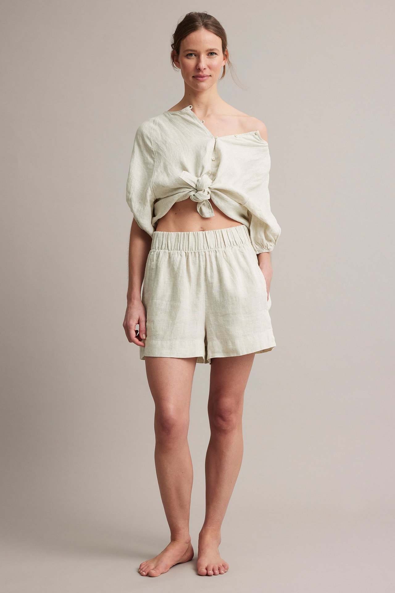 Framifrån: kvinna i beiga linneshorts med resår i midjan och en knuten linneskjorta.