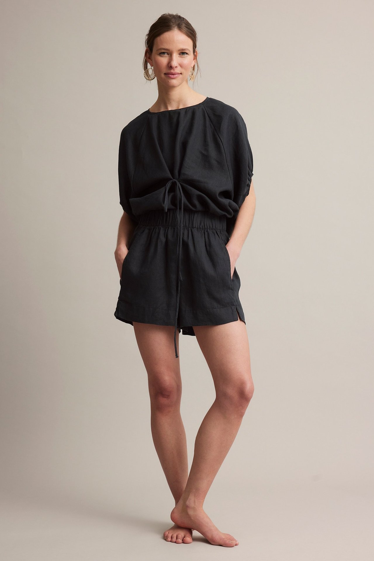 Framifrån: Kvinna i svarta linneshorts, resår i midjan, sidfickor, matchande topp, guldörhängen.