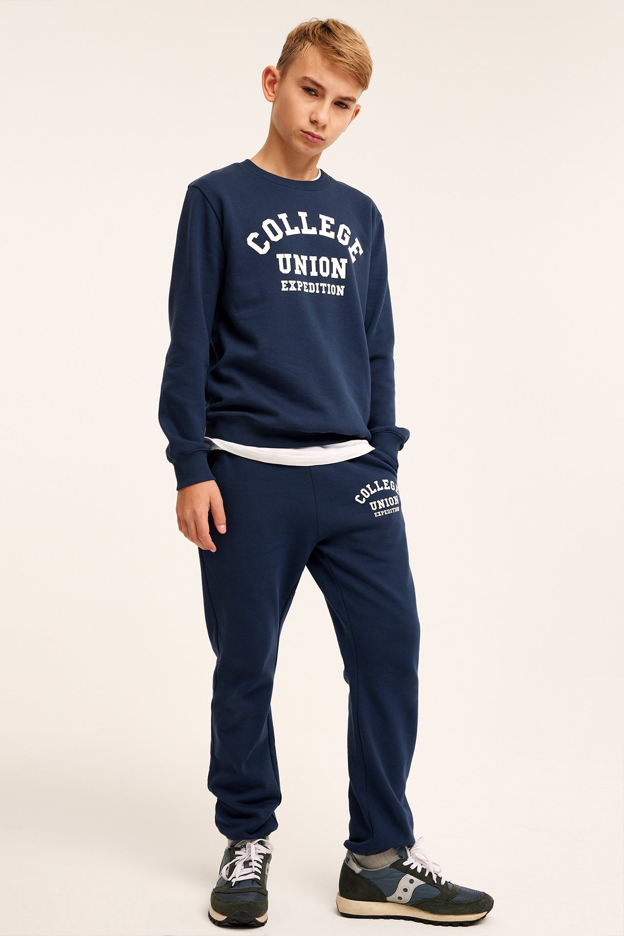 Frontvy av ett barn i mörkblått jogging-set med vitt 'COLLEGE UNION'-tryck, stylat med sneakers.