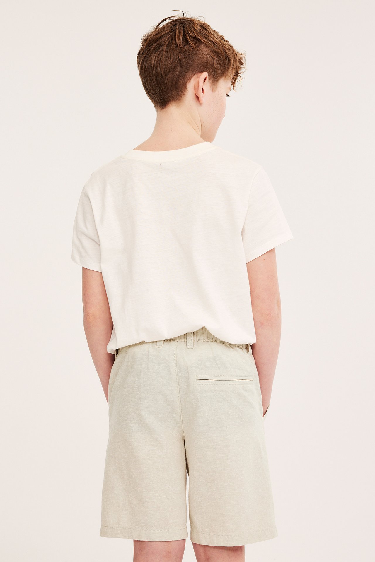 Bakfra: et barn i lysebeige linblanding-shorts med elastisk midje og en lys t-skjorte.