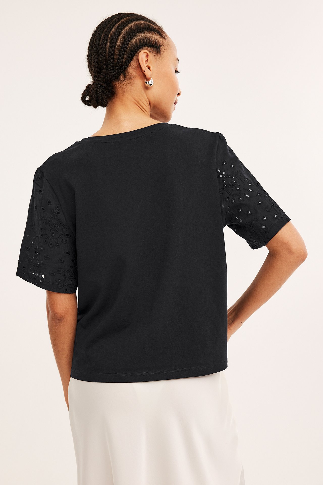 Naisen selkäkuva mustassa puuvilla-T-paidassa, jossa broderie anglaise -hihat, ja valkoisessa hameessa.