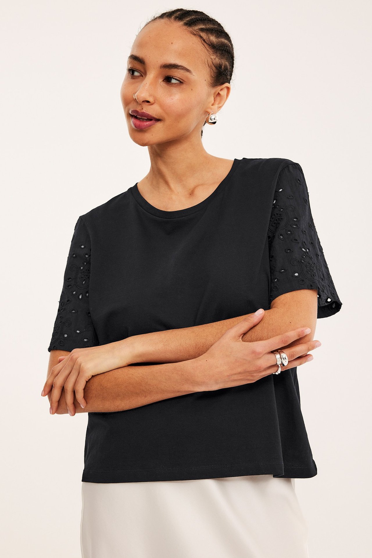 Kobieta w czarnym t-shircie z rękawami broderie anglaise, jasnej spódnicy, srebrnych kolczykach i pierścionkach.