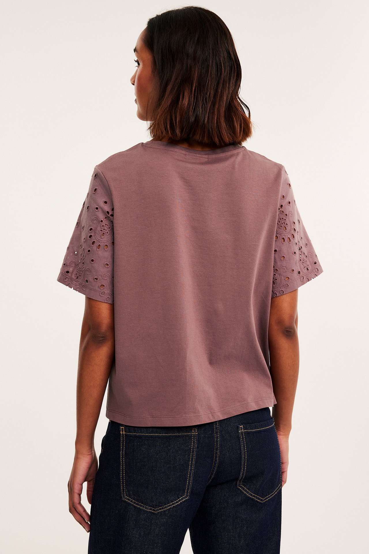 Bakifrån: kvinna i mörkröd bomulls-T-shirt med broderie anglaise-ärmar och mörkblå jeans.