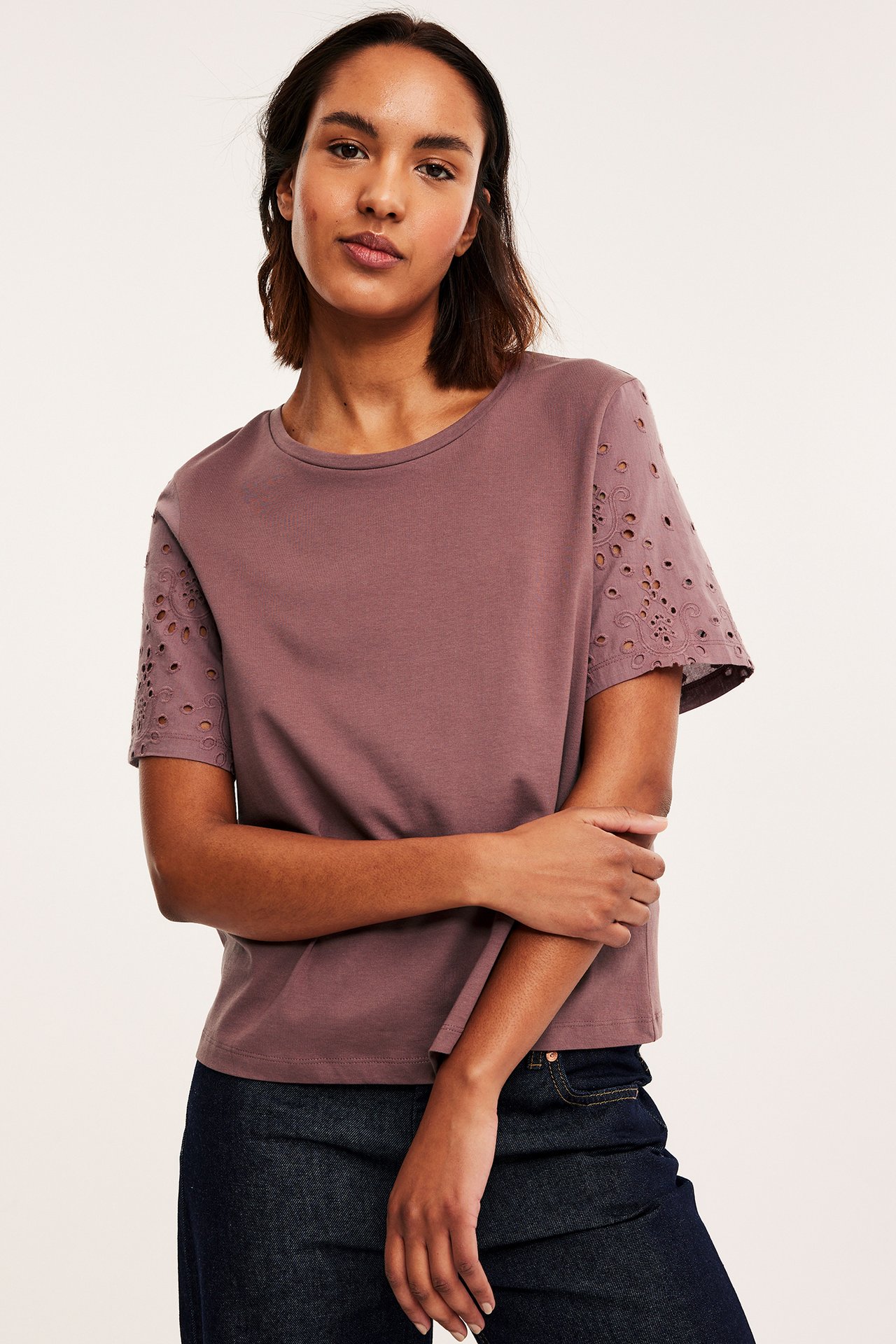 Framifrån, kvinna i mauvefärgad bomulls-T-shirt med broderie anglaise-ärmar och mörkblå jeans.