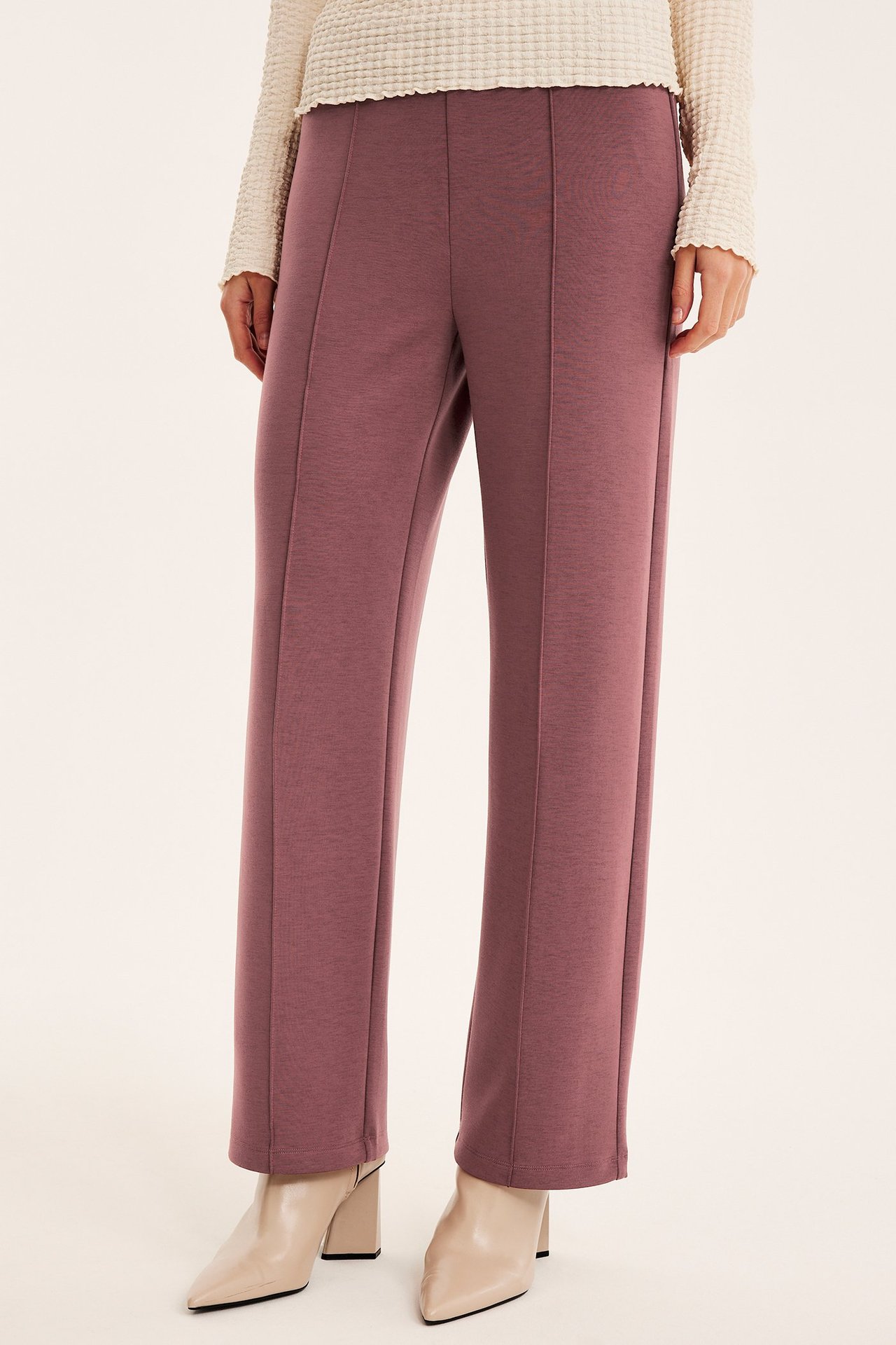 Forfra, kvinne i mørk rosa vide jerseybukser med frontsømmer, en kremfarget topp og beige ankelstøvler.