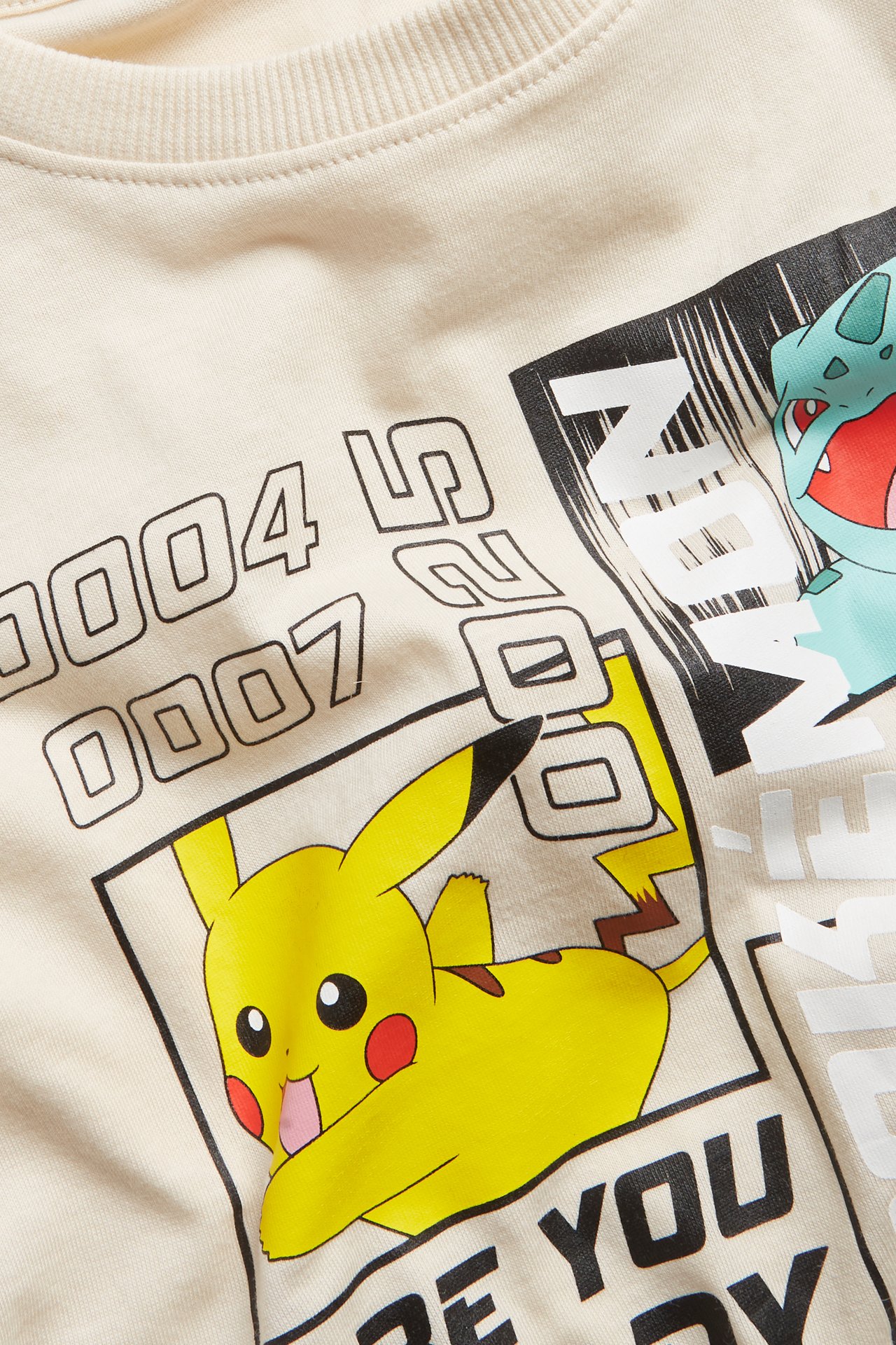 Zbliżenie przodu dziecięcej jasnobeżowej bawełnianej koszulki z dużym nadrukiem Pokémon.