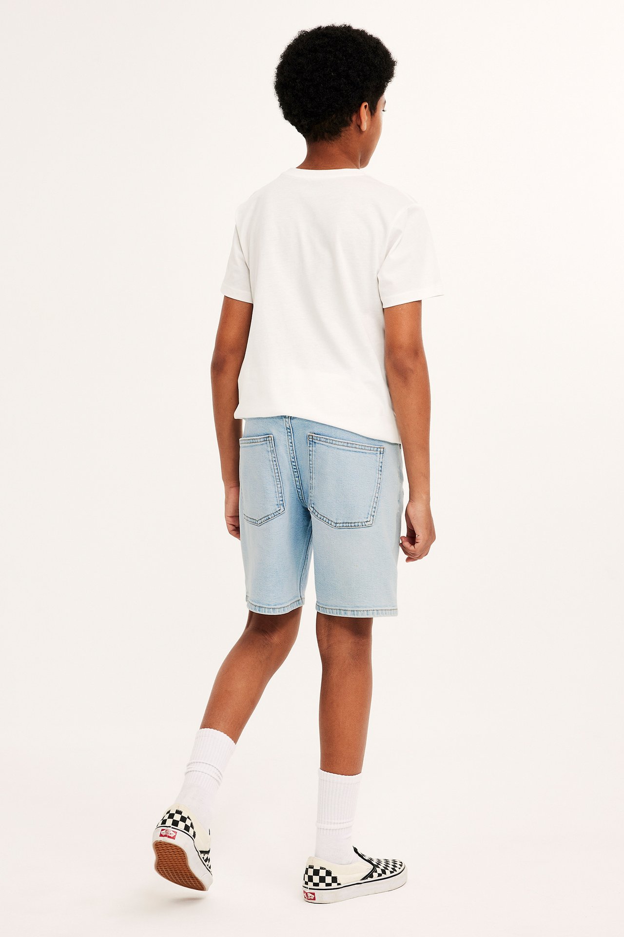 Bakifrån, ett barn i ljusblå stretchiga jeansshorts, vit t-shirt, vita strumpor och rutiga slip-on skor.