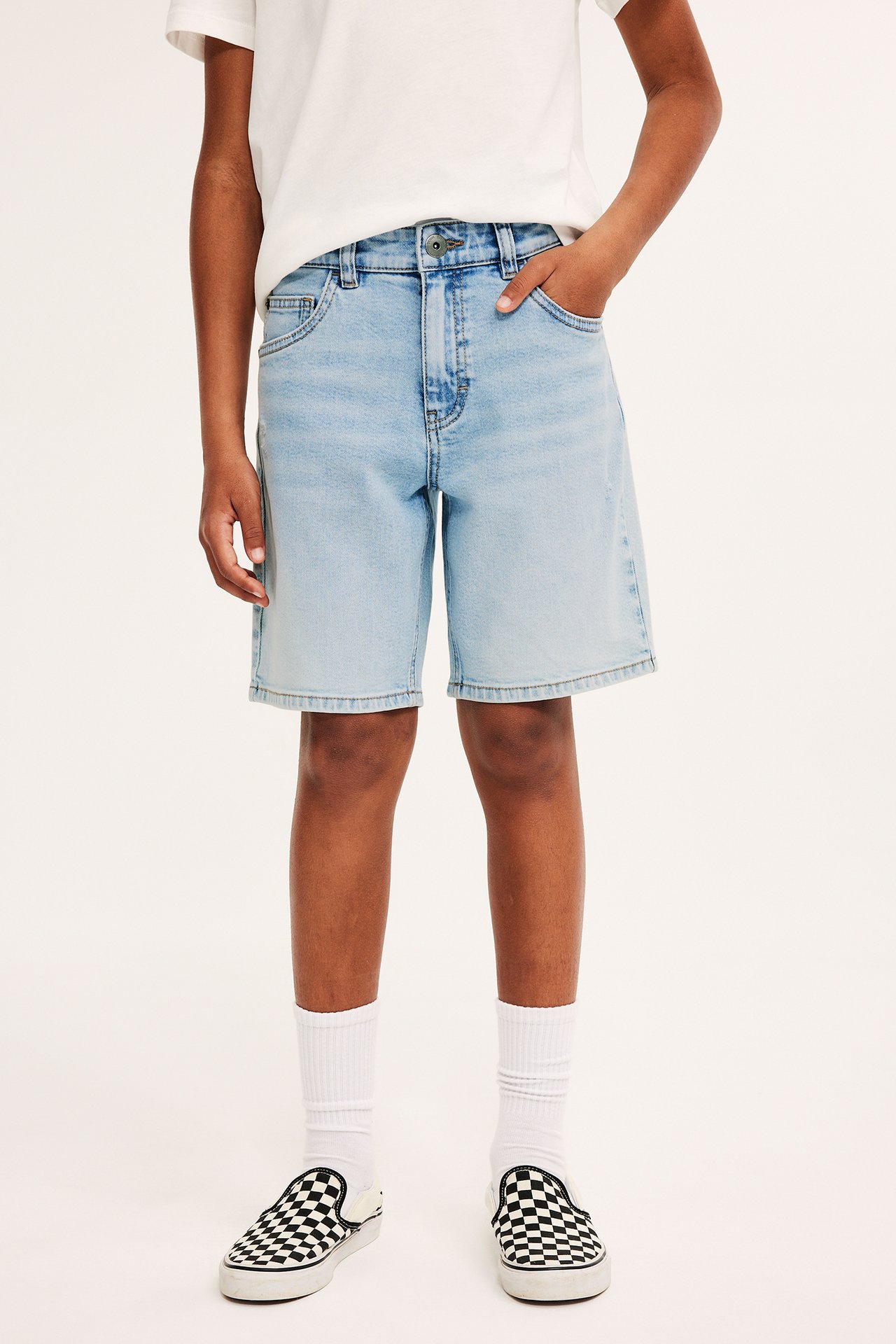 Framifrån: Barn bär ljusa stretchjeansshorts, vit t-shirt, vita strumpor och rutiga slip-on skor.