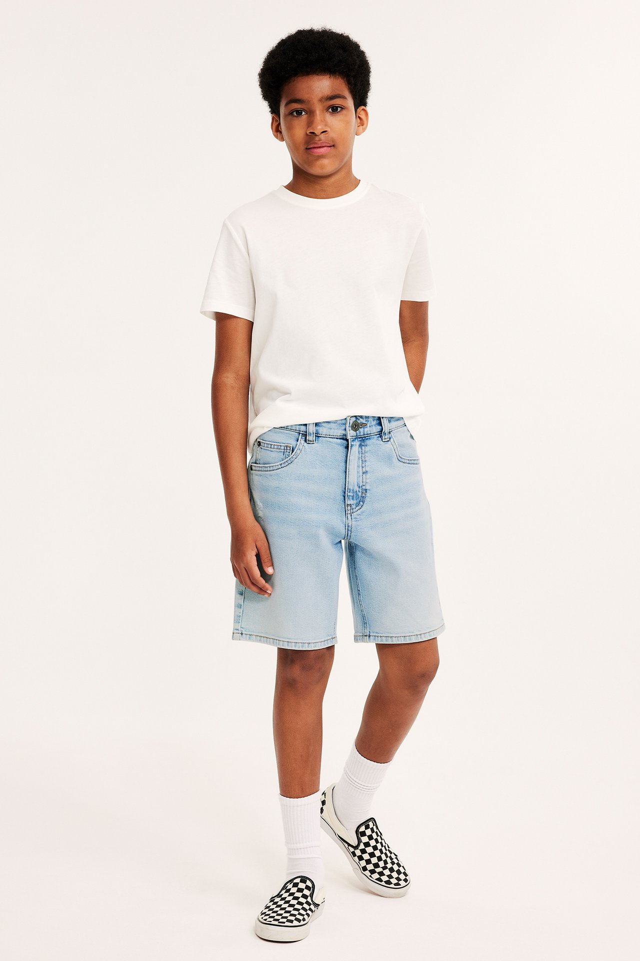 Framifrån: ett barn i ljusblå denimshorts, vit t-shirt, vita strumpor och rutiga slip-on-skor.