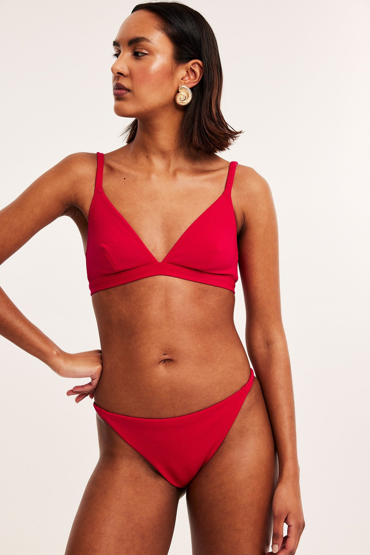 Fram-sidovy av en kvinna i röd strukturerad triangel bikini-bh, matchande trosa och guldspiralörhängen.