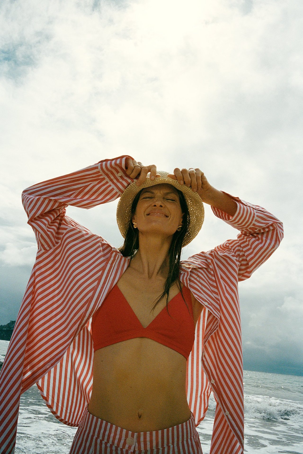 Kobieta w czerwonym teksturowanym biustonoszu bikini, czerwono-białej koszuli w paski, pasujących spodniach, trzyma kapelusz. Przód.