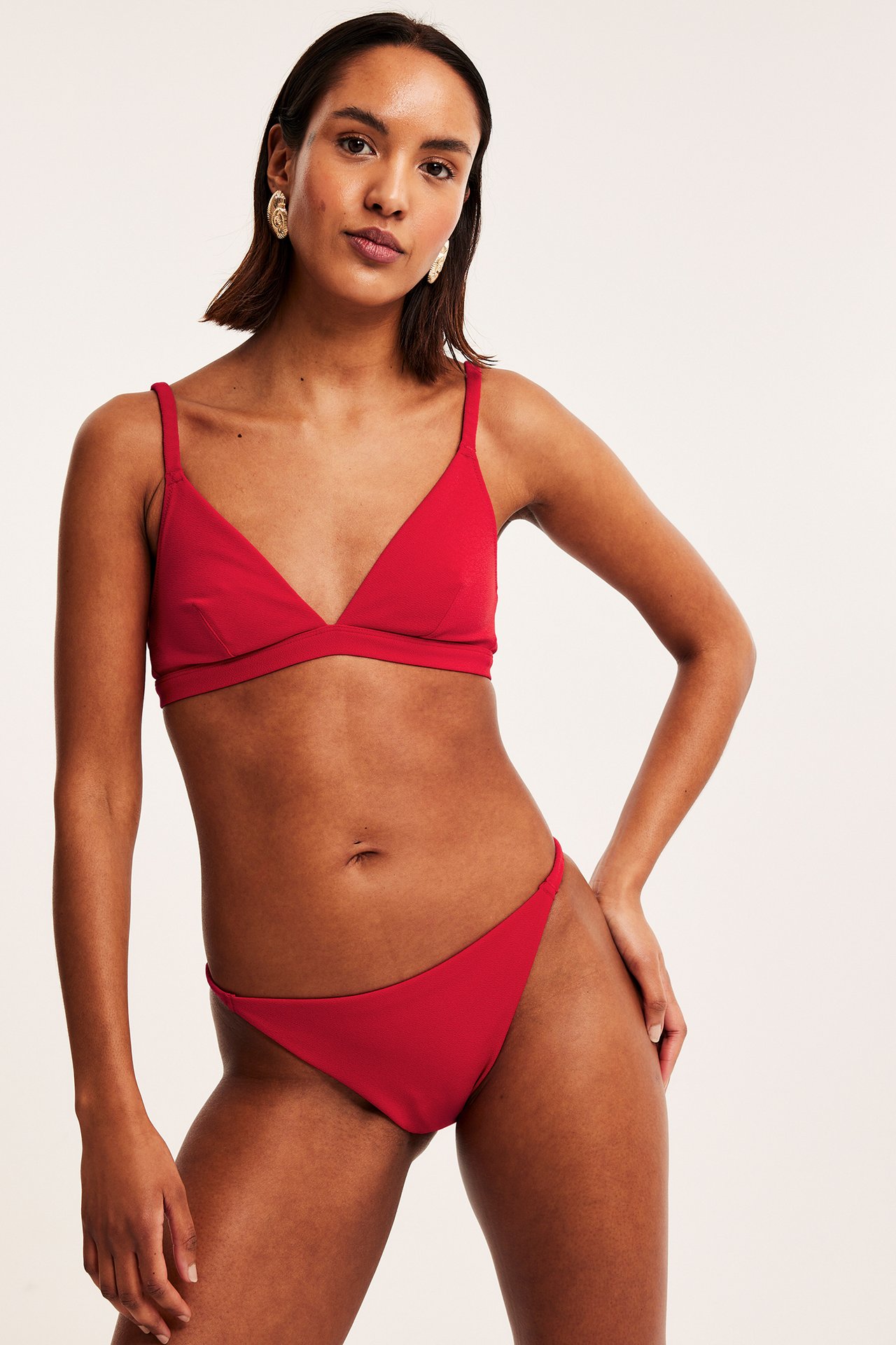 Edestä kuvattu nainen yllään punaiset kuvioidut brazilian-bikinihousut, yhteensopiva kolmio-bikinitoppi ja kultaiset korvakorut.