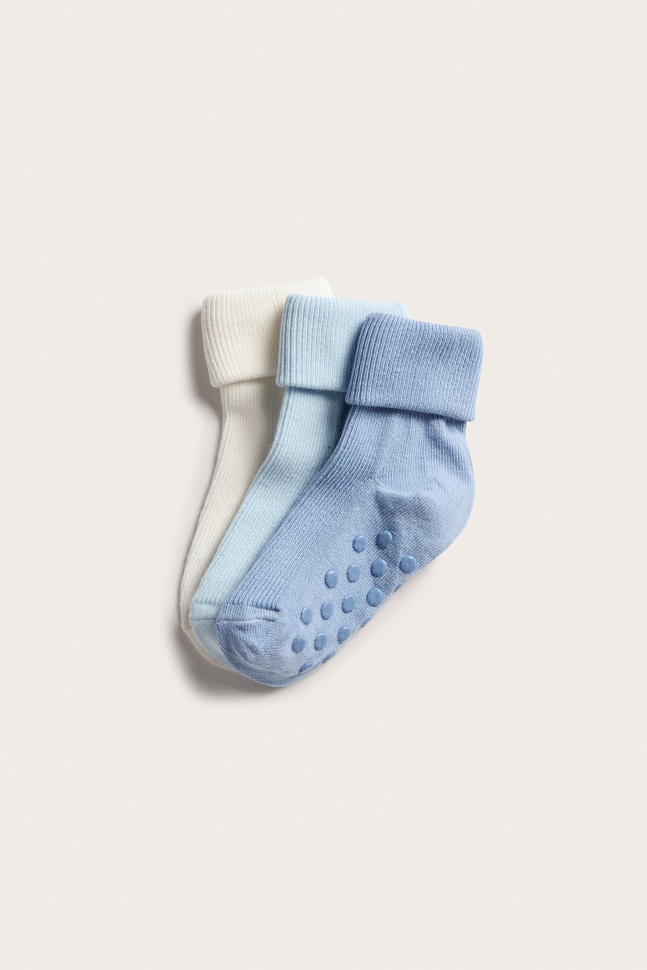 Topputsikt av tre babysokker: kremhvit, lyseblå og blå, med brettede mansjetter og antiskli såler.
