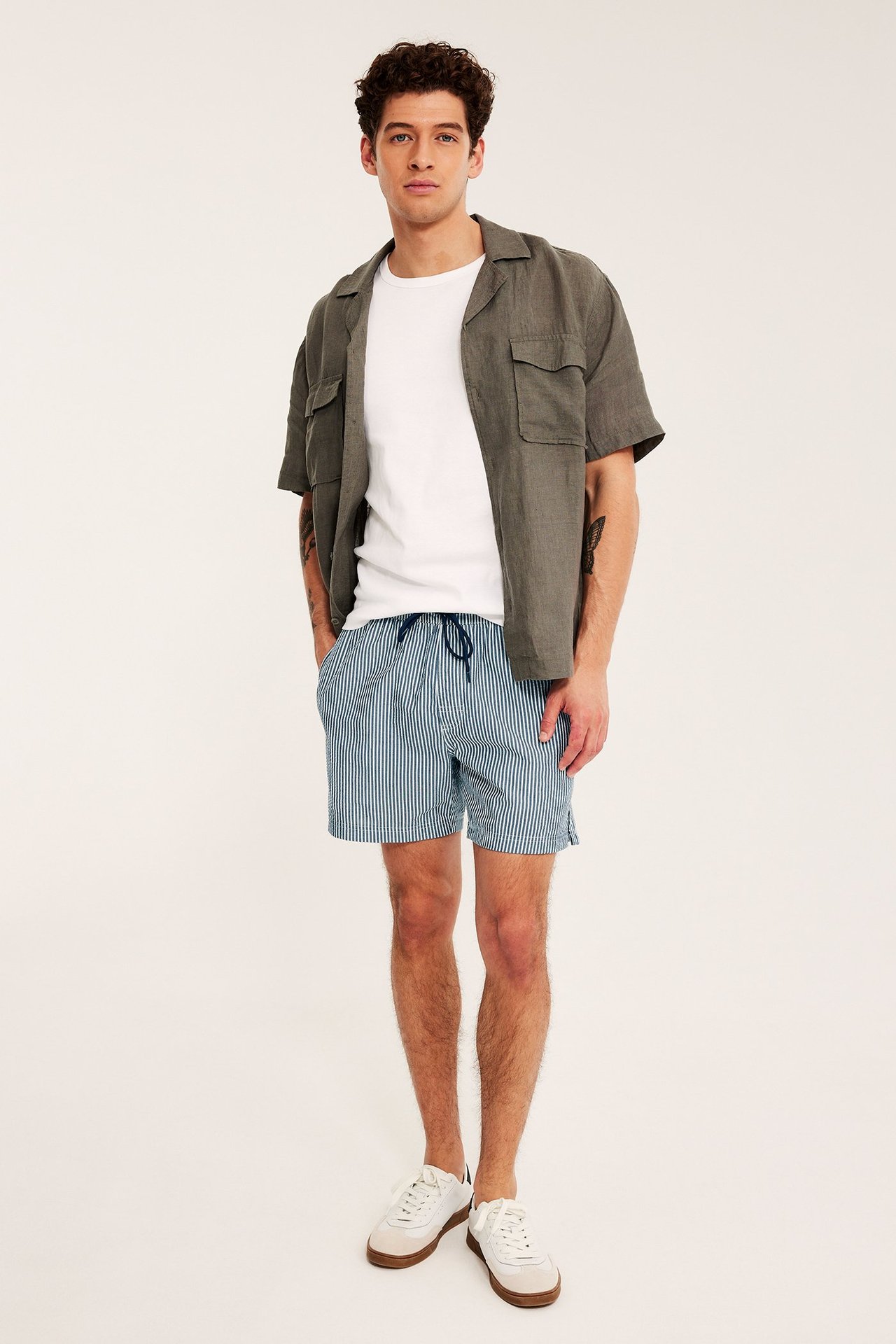Etunäkymä: mies raidallisissa tummansinisissä seersucker-uimashortseissa, valkoinen t-paita ja avoin oliivinvihreä lyhythihainen paita.