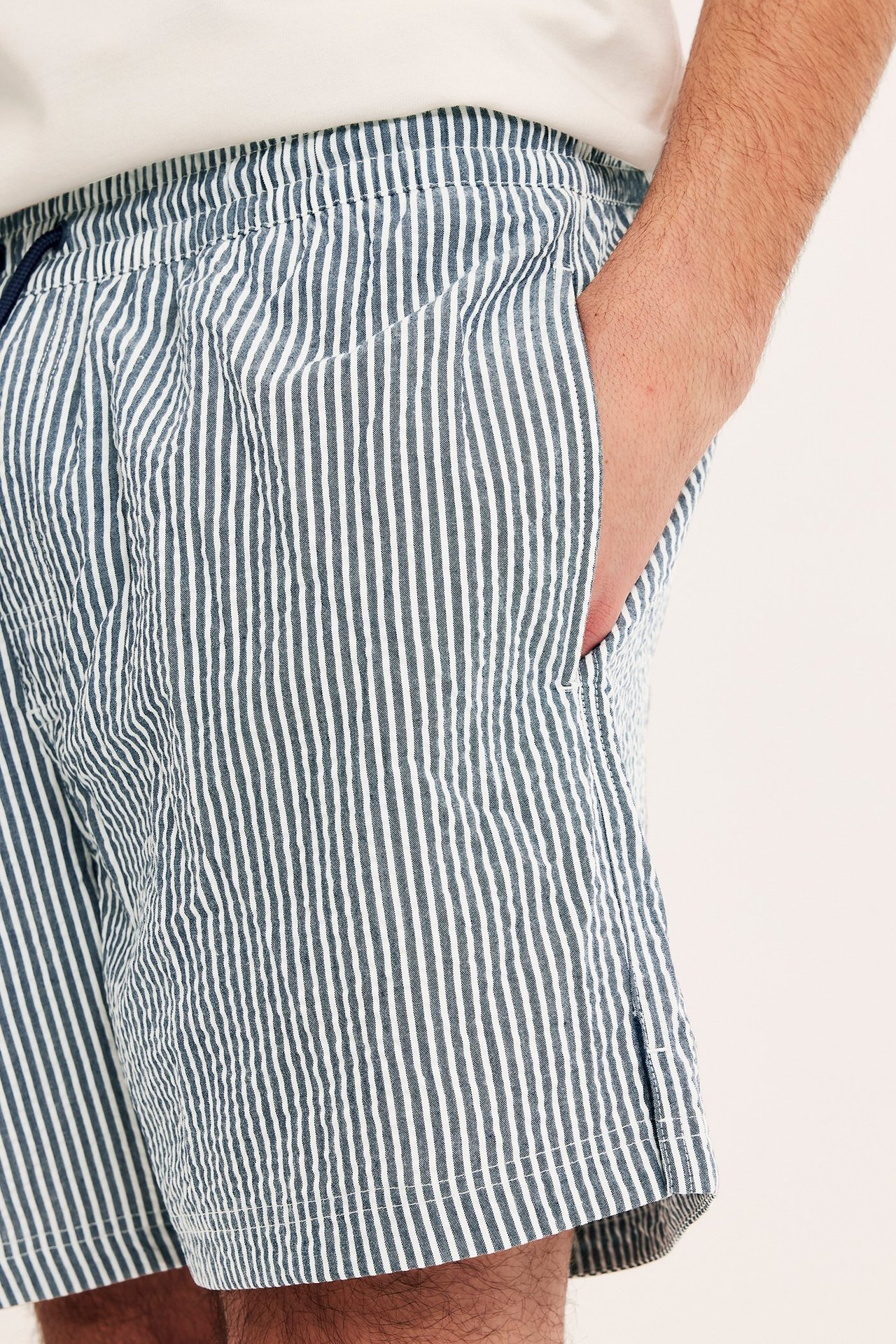 Lähikuva, sivukuva miehestä yllään tummansiniset-valkoiset raidalliset seersucker-uimashortsit, käsi taskussa.