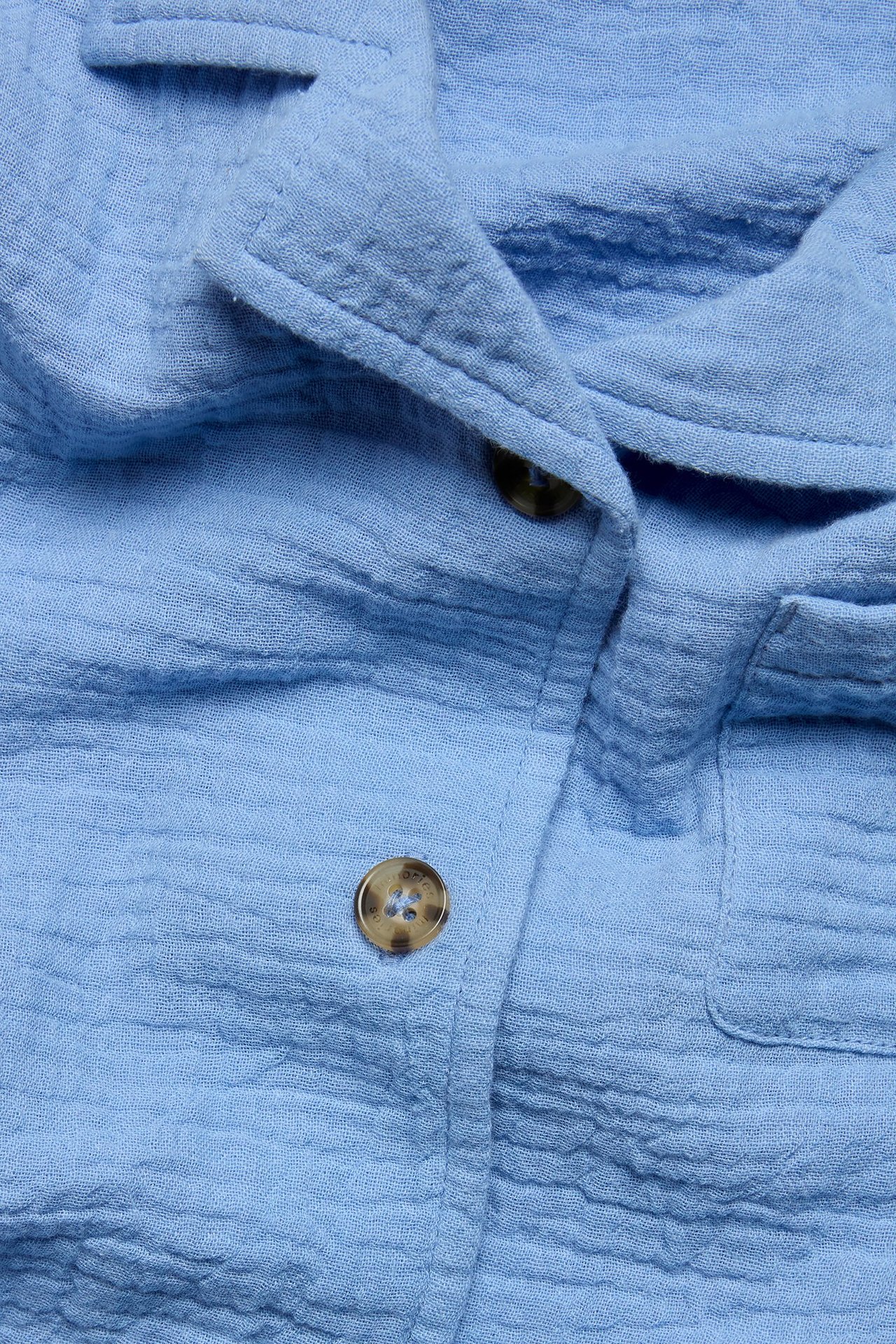 Zbliżenie na jasnoniebieską koszulę dziecięcą z muślinu, z brązowymi guzikami i kieszenią.