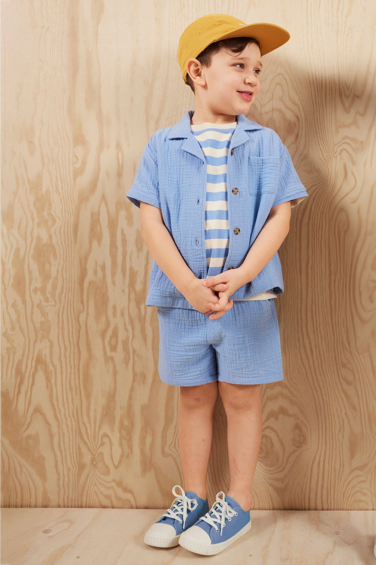 Widok z przodu dziecka w jasnoniebieskiej koszuli z krótkim rękawem, pasujących szortach, pasiastym t-shircie, żółtej czapce i niebieskich trampkach.