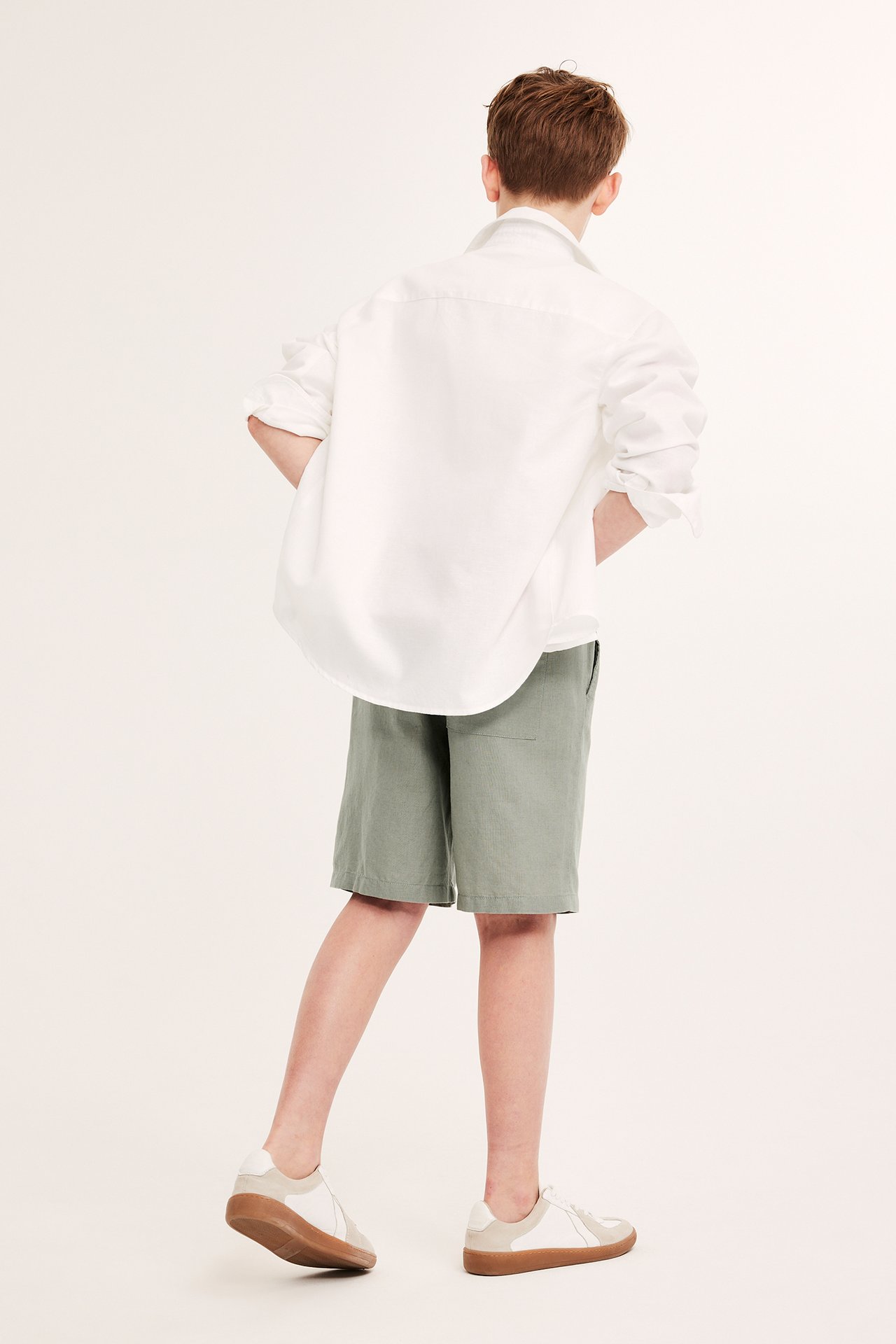Bakifrån: Kid i gröna linneshorts, vit långärmad skjorta och vita och beiga sneakers.