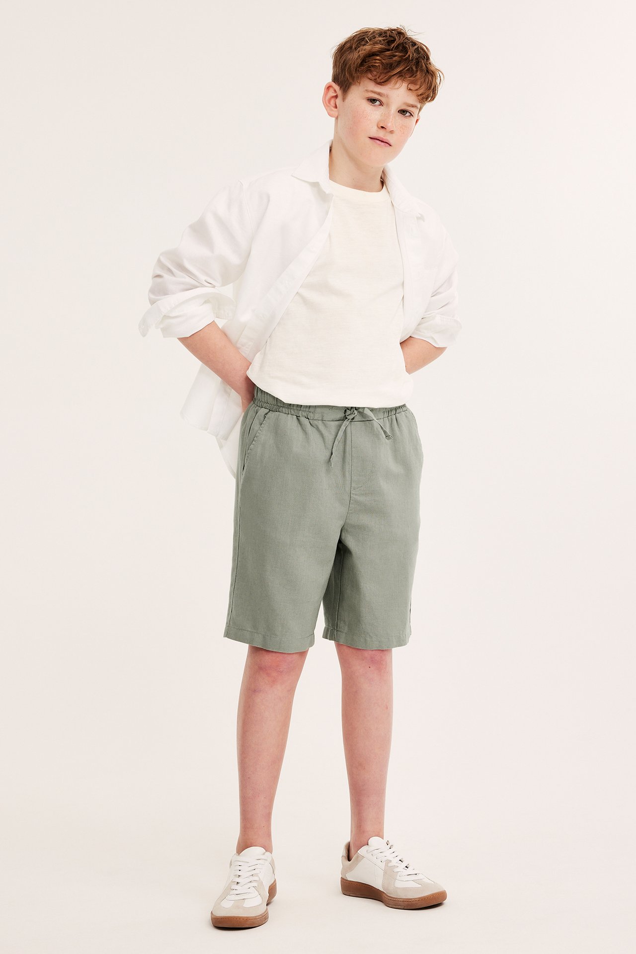 Framifrån: Barn i gröna linneshorts, vit t-shirt, öppen vit skjorta och vita sneakers.
