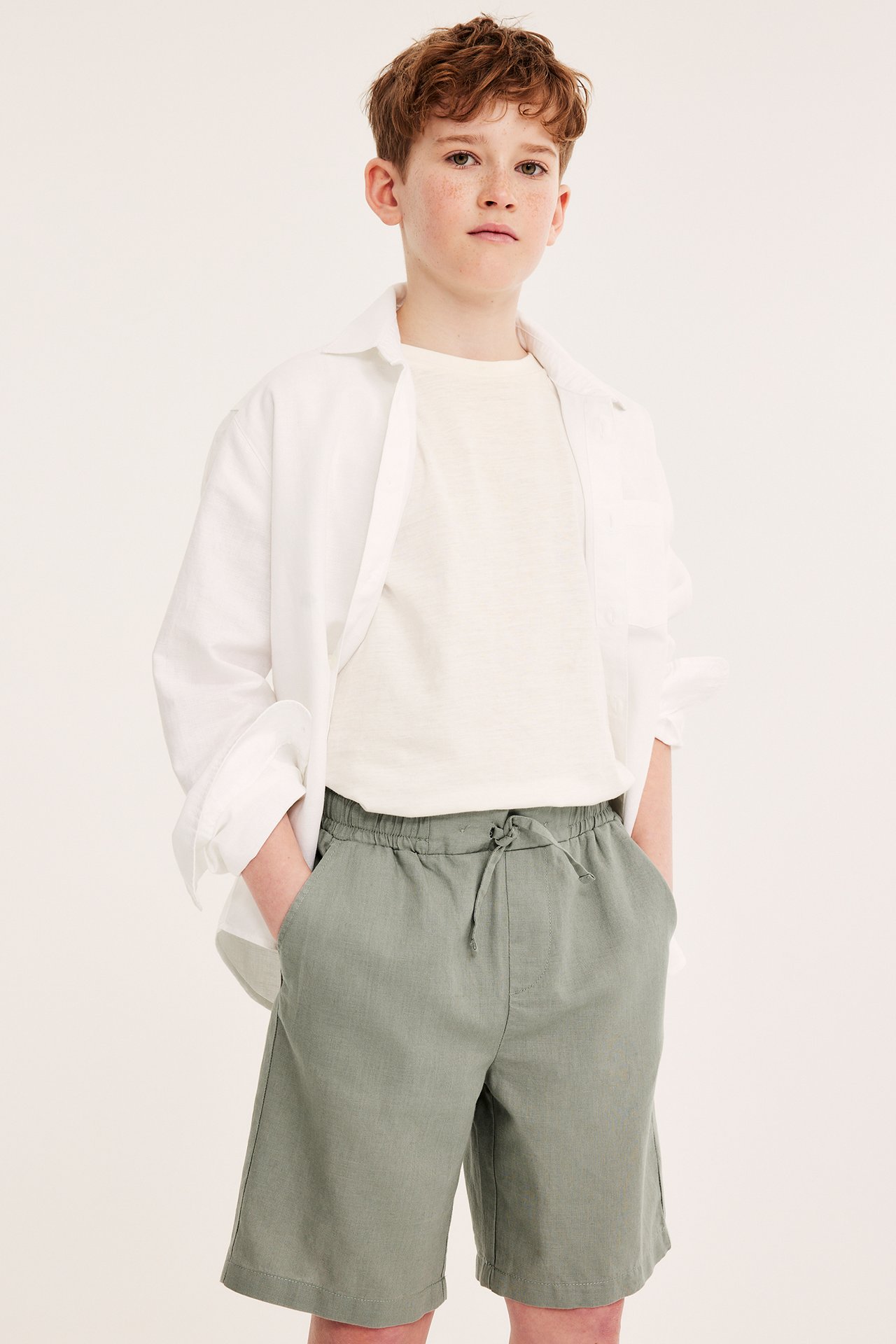 Frontvy av ett barn i gröna linneshorts med dragsko, ljusbeige t-shirt och öppen vit skjorta.