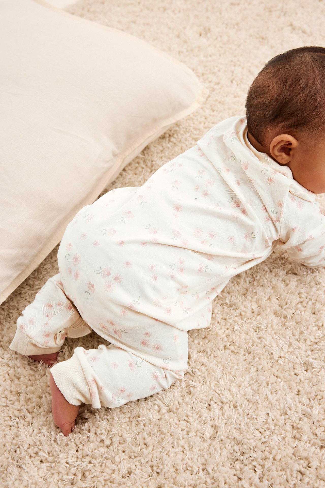 Ylhäältä otettu takakuva ryömivästä vauvasta, yllään luonnonvalkoinen kuviollinen pyjama vaaleanpunaisilla kukilla.