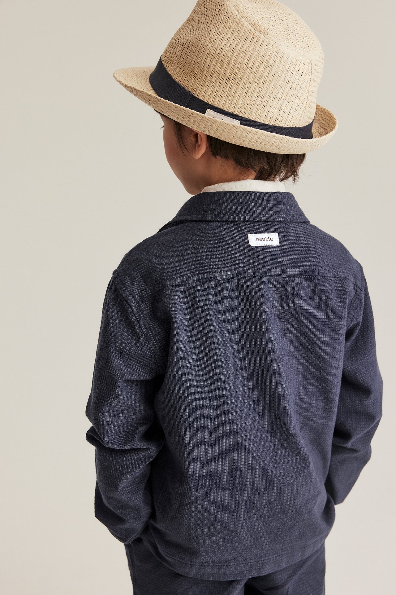 Bakfra: et barn i mørkeblå chambray-overshirt og en beige stråhatt med mørkt bånd.