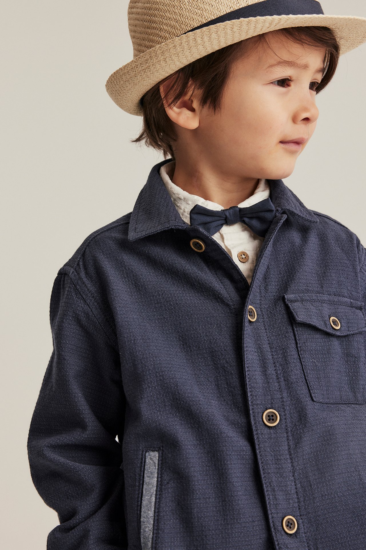 Barn har på seg en mørkeblå chambray-overskjorte, hvit skjorte, mørkeblå sløyfe og beige stråhatt.