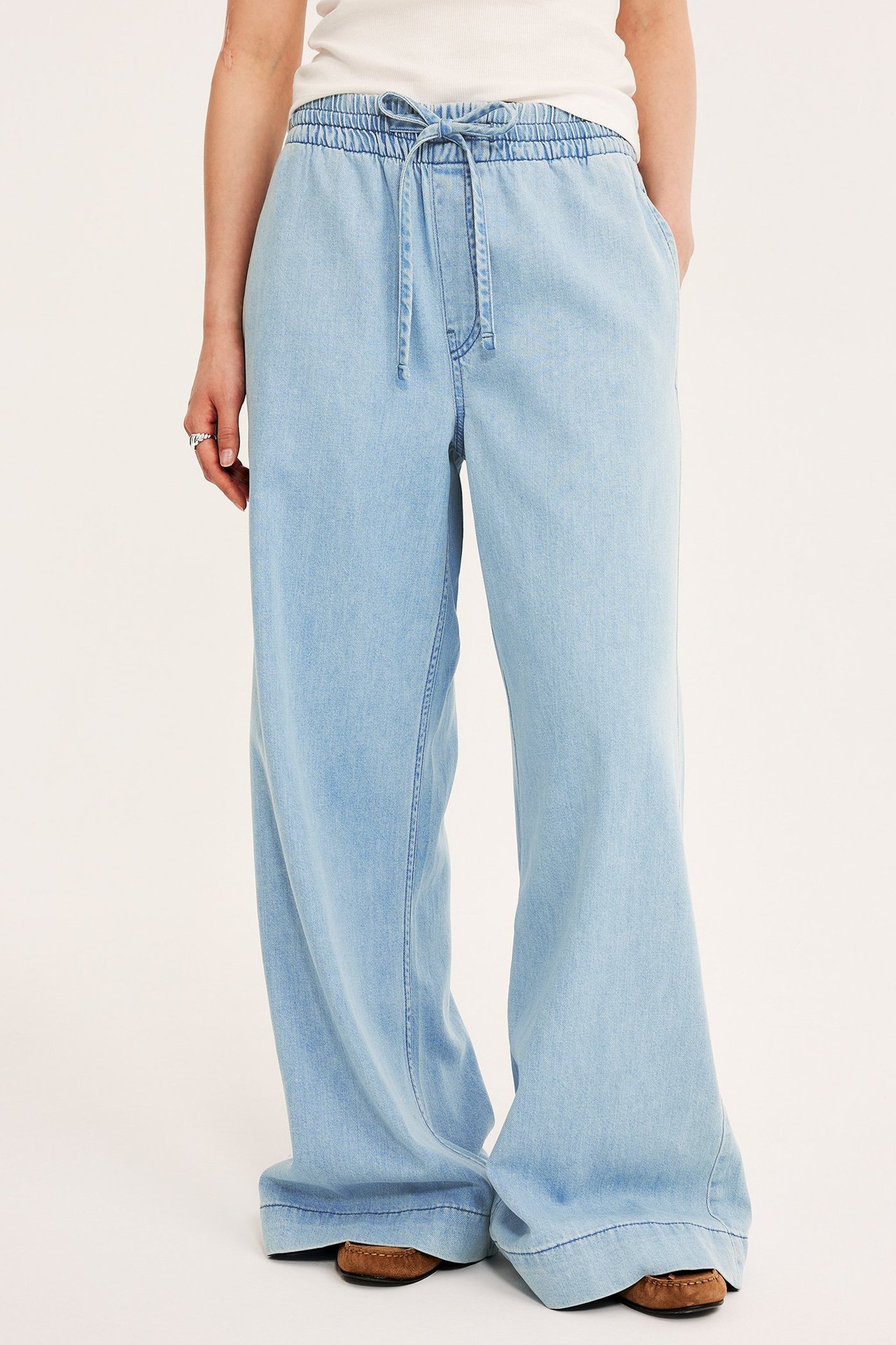 Framifrån: Kvinna bär ljusblå vida pull-on jeans med dragsko, vit topp, bruna skor.
