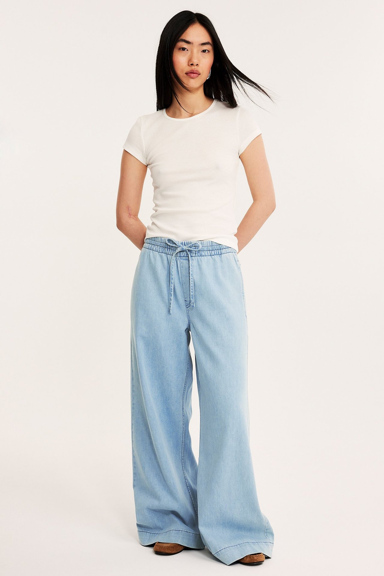 Frontvy av en kvinna i ljusblå vida pull-on jeans med dragsko i midjan och en vit t-shirt.