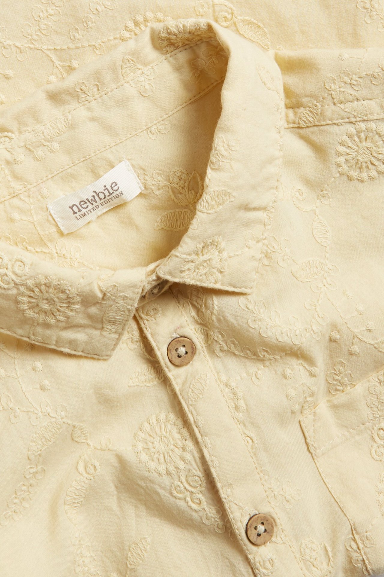 Zbliżenie żółtej bawełnianej koszuli z krótkim rękawem, z kwiatowym haftem i drewnianymi guzikami.