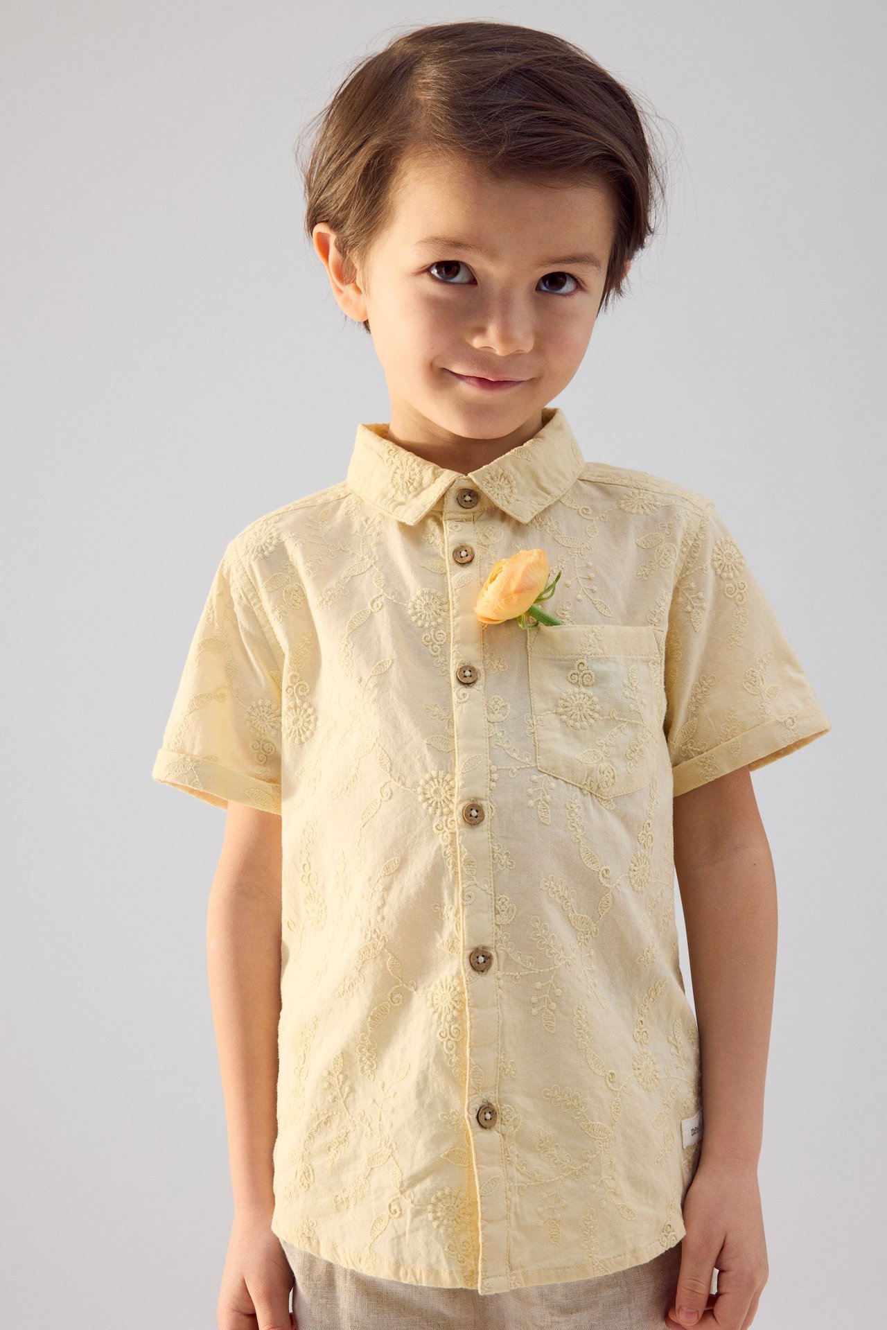 Widok z przodu dziecka w żółtej bawełnianej koszuli z krótkim rękawem, haftem kwiatowym i kwiatem w kieszeni.
