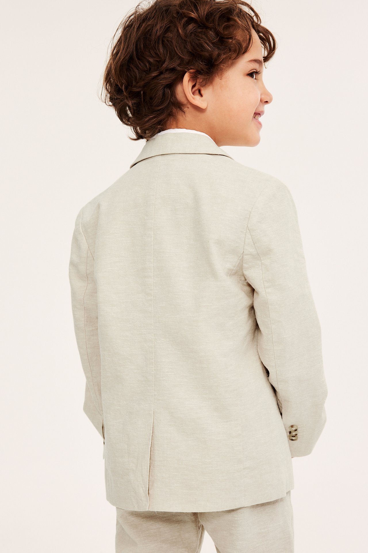 Bakfra, et barn i lys beige linblanding blazer over hvit skjorte, med matchende bukser.