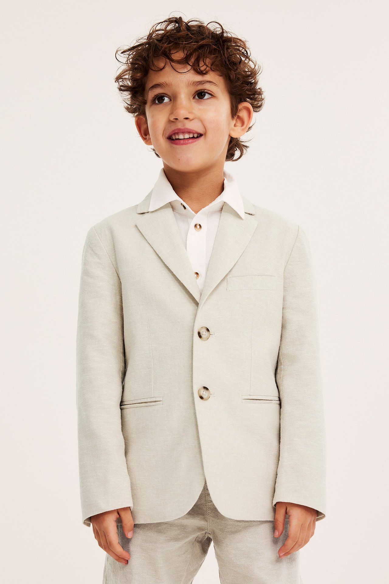 Edestä hymyilevä lapsi vaaleanbeigessä pellavasekoitekavajassa, valkoisessa paidassa ja yhteensopivissa housuissa.