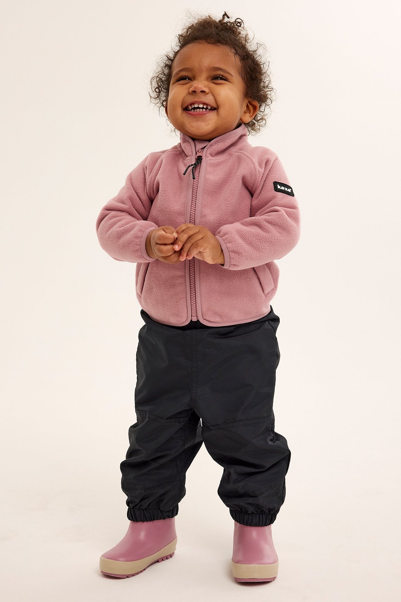 Edestä hymyilevä vauva mustissa kuorihousuissa, vaaleanpunaisessa fleecetakissa ja vaaleanpunaisissa kumisaappaissa.