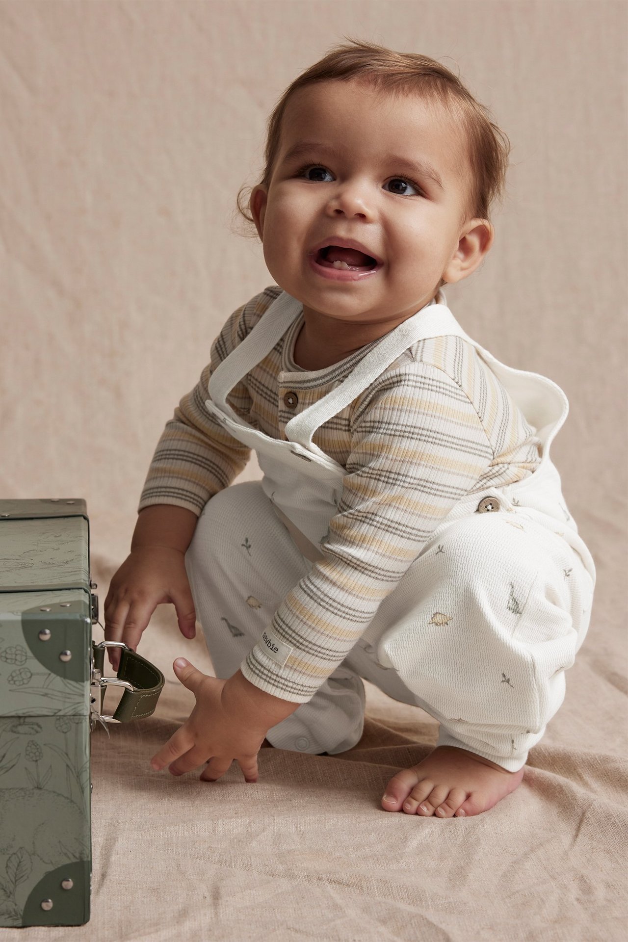 Framifrån: baby i offwhite hängselbyxor med dinosauriemönster över randig långärmad tröja, nära grön låda.