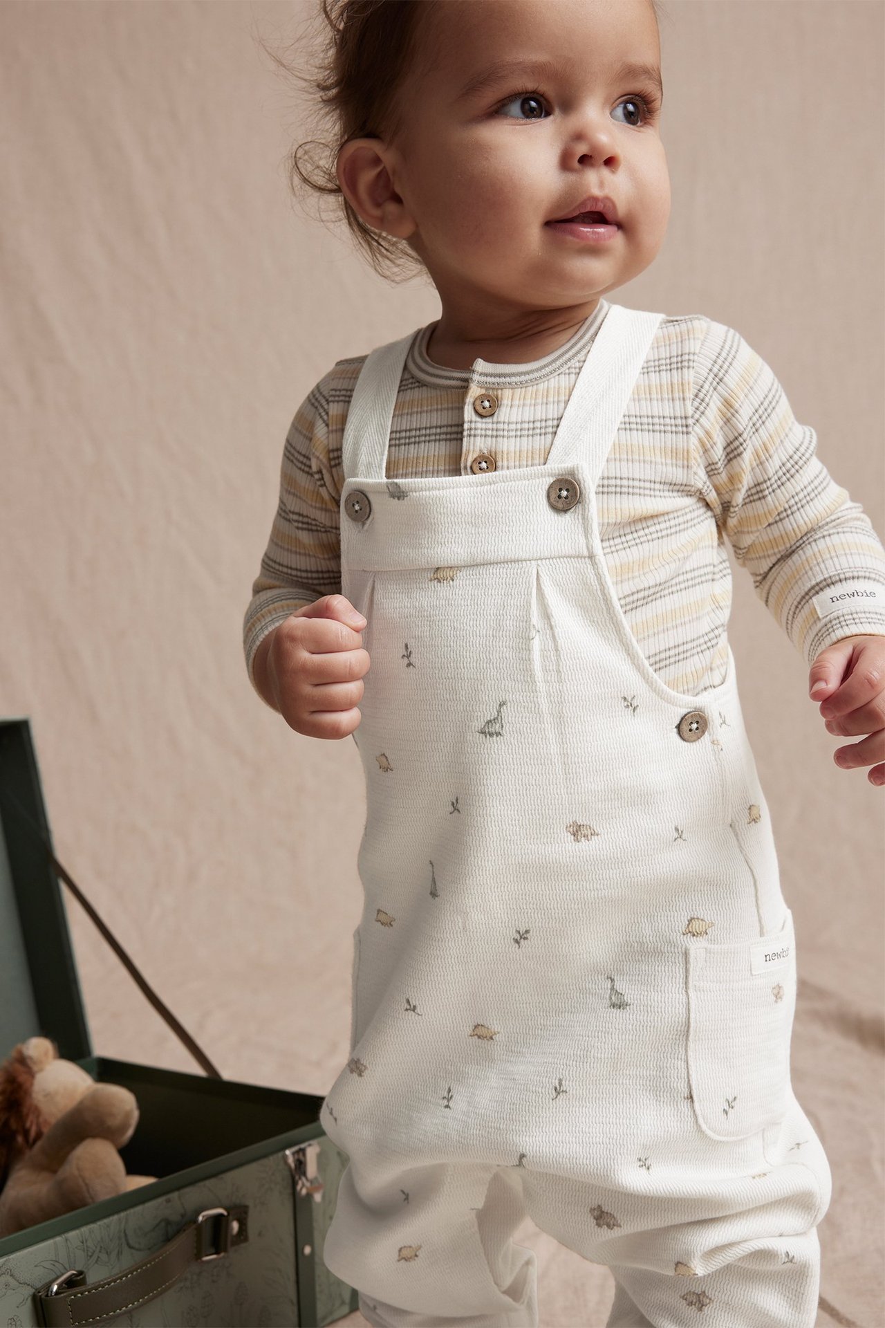 Frontvy av baby i offwhite hängselbyxor med dinosauriemönster över en randig långärmad tröja.