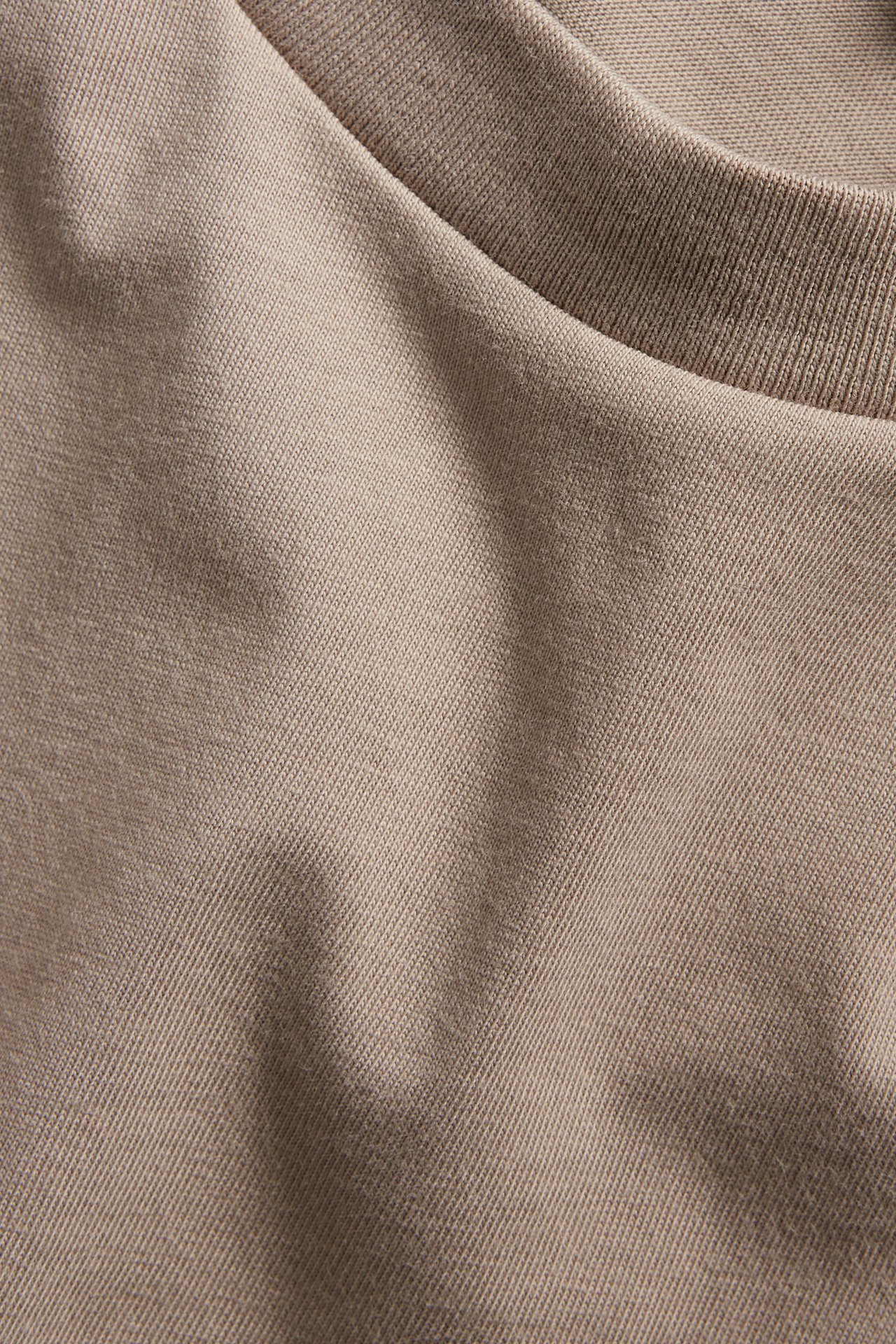 Nærbilde av en mørk beige oversized bomulls-T-skjorte, viser ensfarget stoff og ribbet krage.