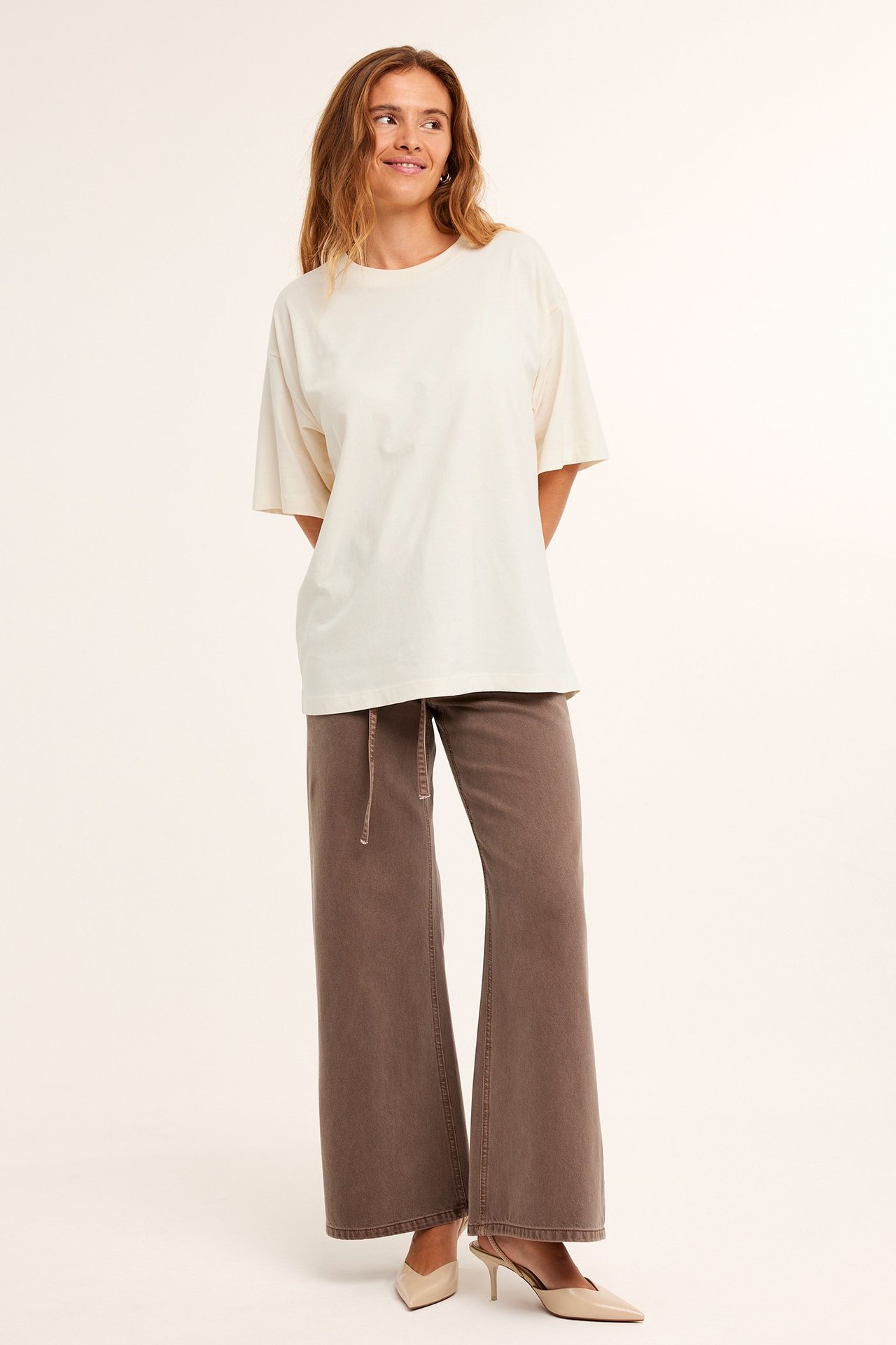 Frontvisning av en kvinne i en offwhite oversized bomulls-T-skjorte, brune vide bukser og beige hæler.