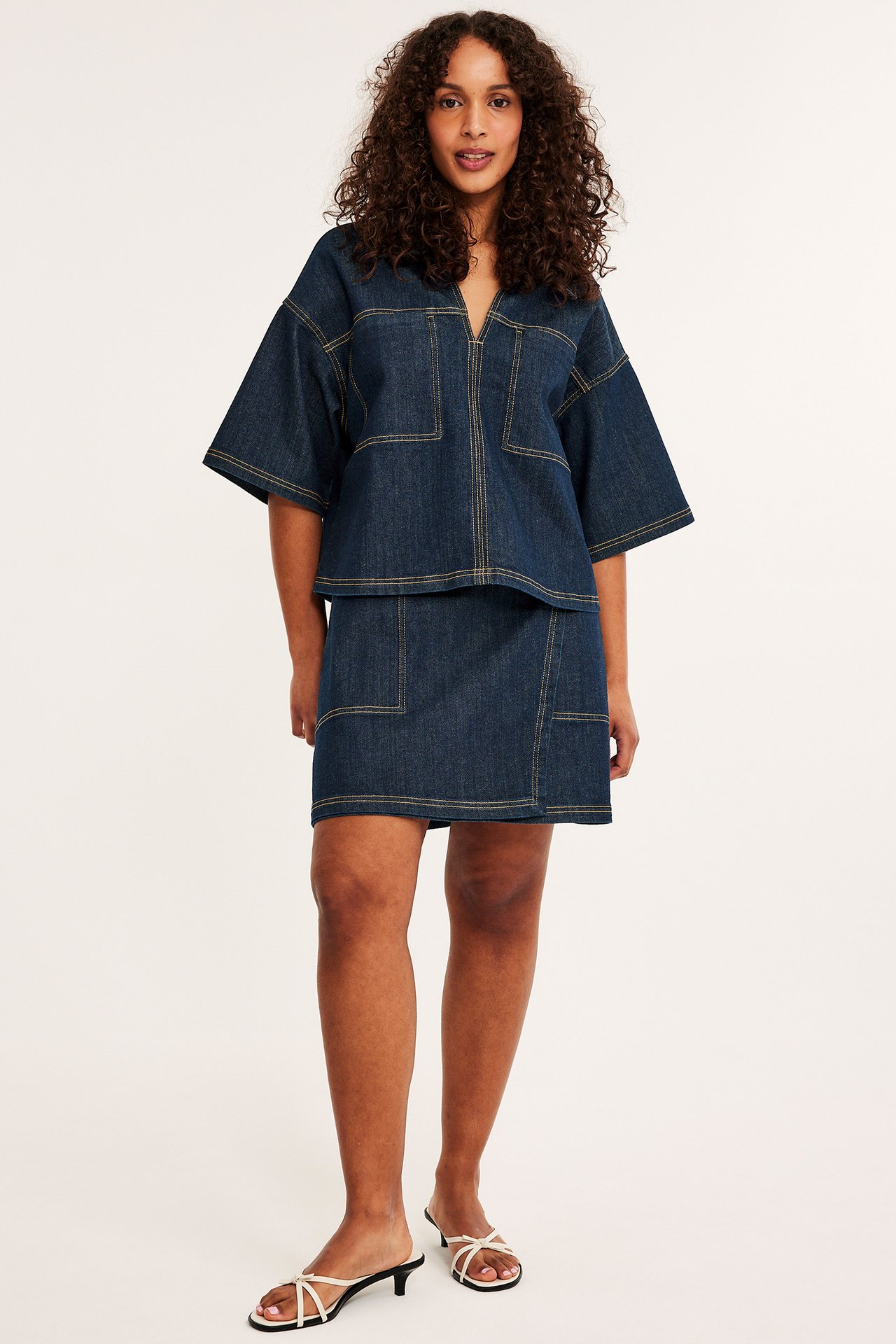 Edestä: Nainen tummassa denim-kietaisuminihameessa taskuilla, yhteensopiva toppi ja valkoiset remmisandaalit.