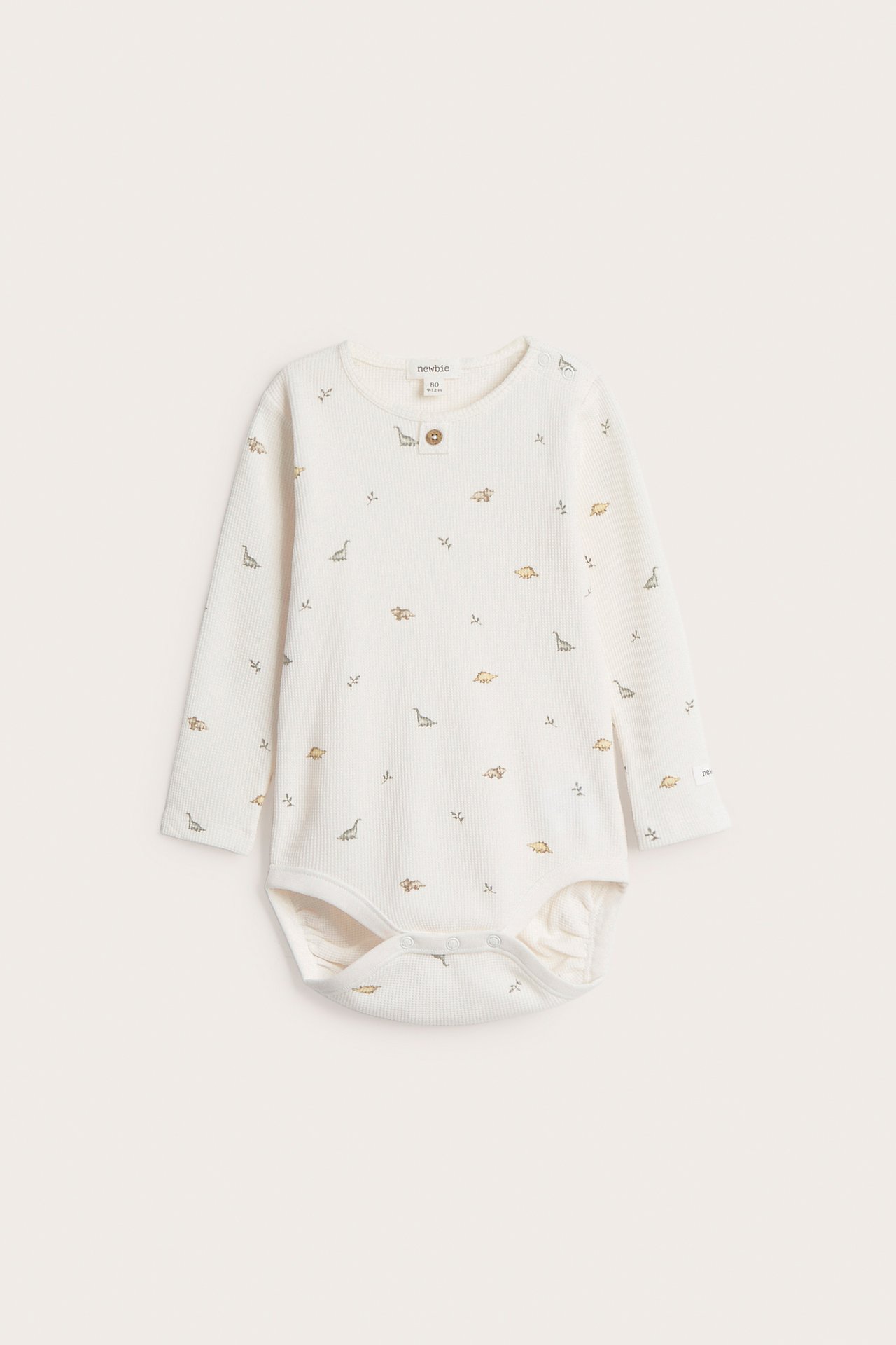 Frontvy av en offwhite våfflad babybody med långa ärmar, dinosauriemönster och tryckknappar.