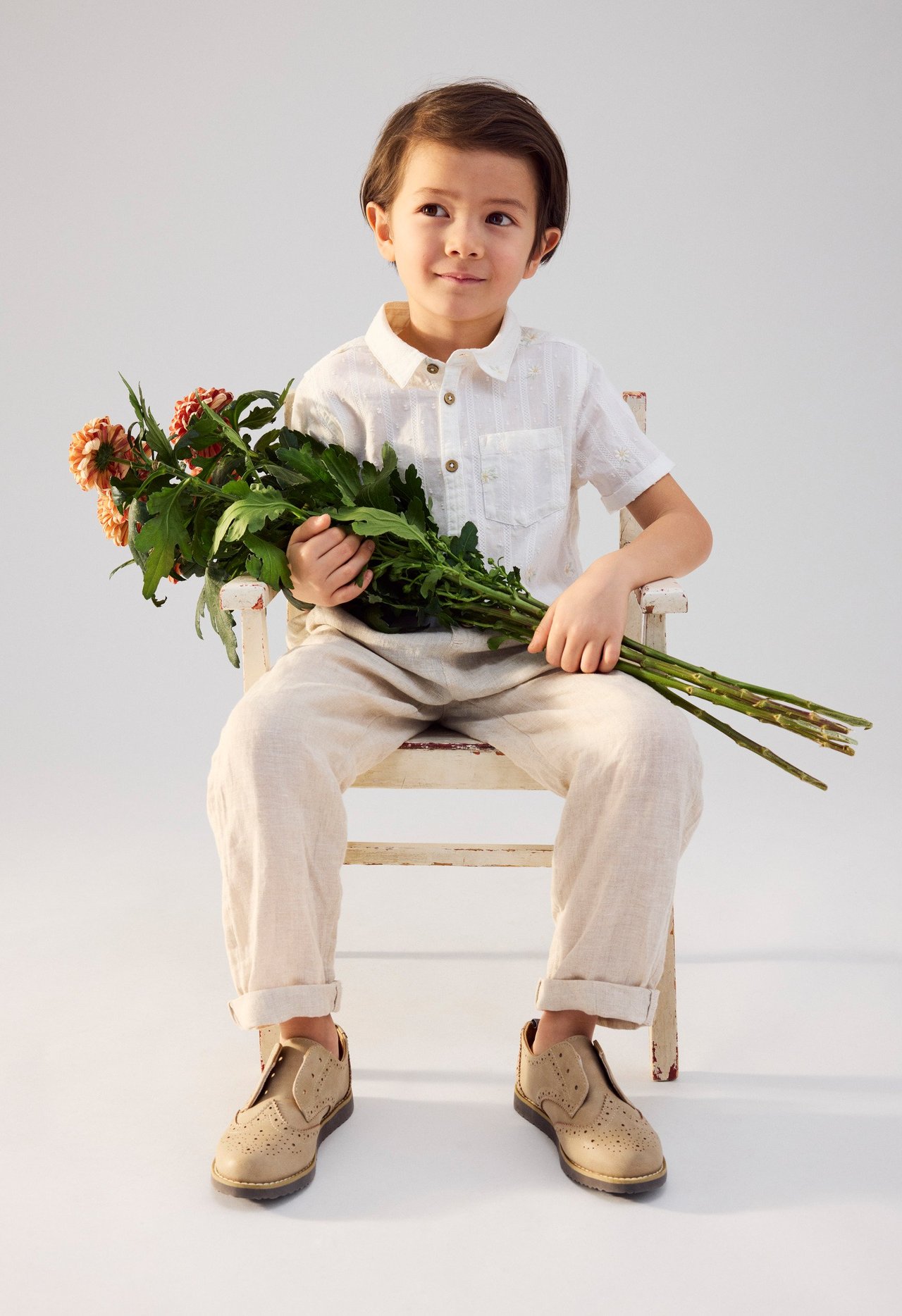 Widok z przodu dziecka w kremowej koszuli bawełnianej w kwiaty, beżowych spodniach i beżowych butach brogues, trzymającego kwiaty.