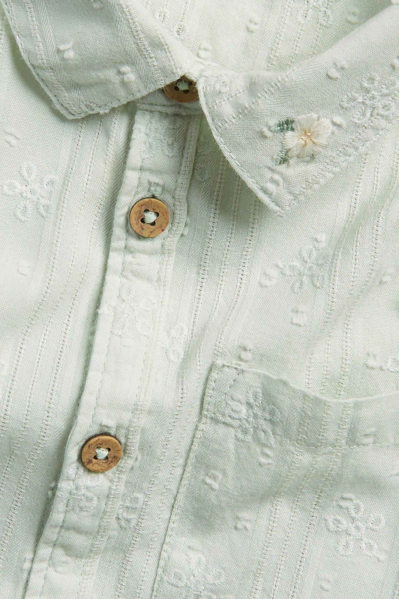 Zbliżenie jasnozielonej bawełnianej koszuli z krótkim rękawem, z haftowanymi detalami, kołnierzykiem i drewnianymi guzikami.